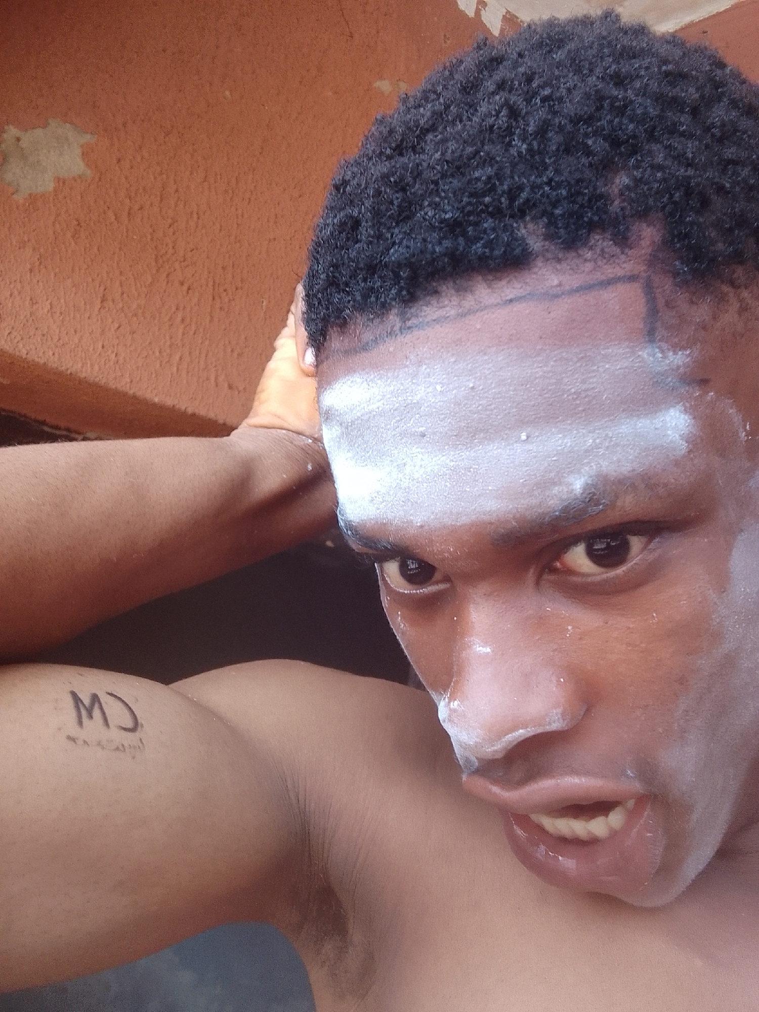 A close-up, selfie-style shot shows a dark-skinned man with short, dark hair. He has white powder or cream covering his forehead and nose. The tattoo 'MJ' can be seen on his left arm. The background appears to be an orange wall. This image was taken in Nkpor, Nigeria. The subject's face is tense, and his mouth is slightly open.