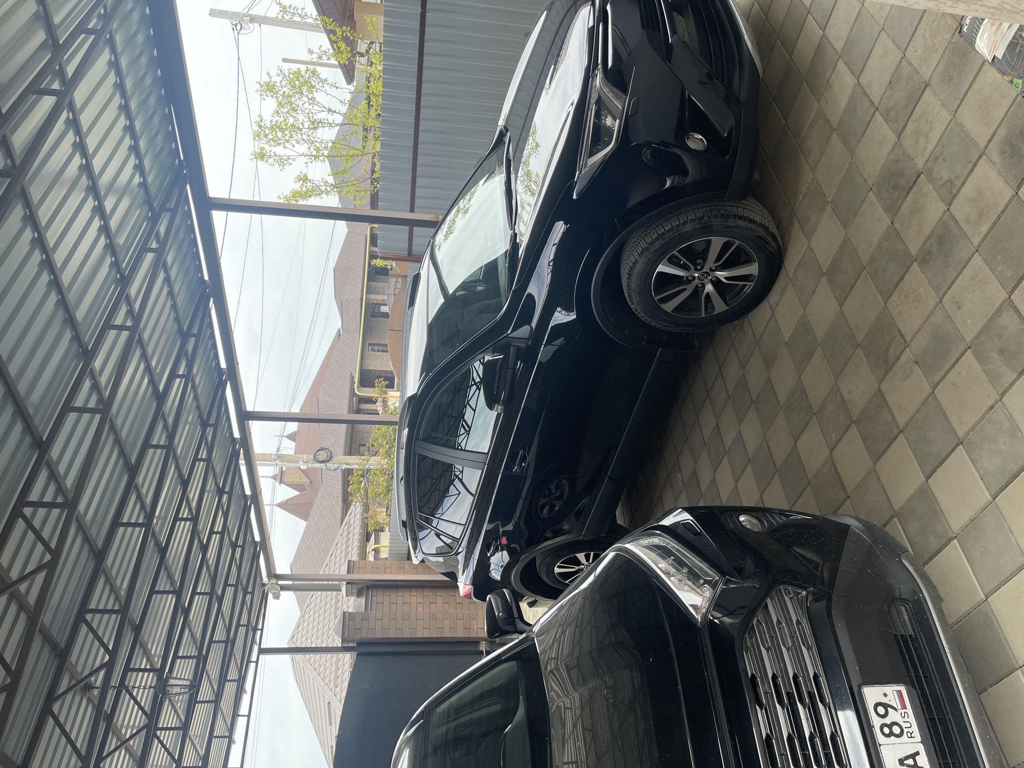 The image shows two black cars parked under a metal awning. The car closer to the camera has a Russian license plate with the number "89". The surrounding environment appears to be a residential area in Yessentukskaya, Russia, as indicated by the location information. The ground is covered in gray and white checkered tiles. A house and a fence are visible in the background.