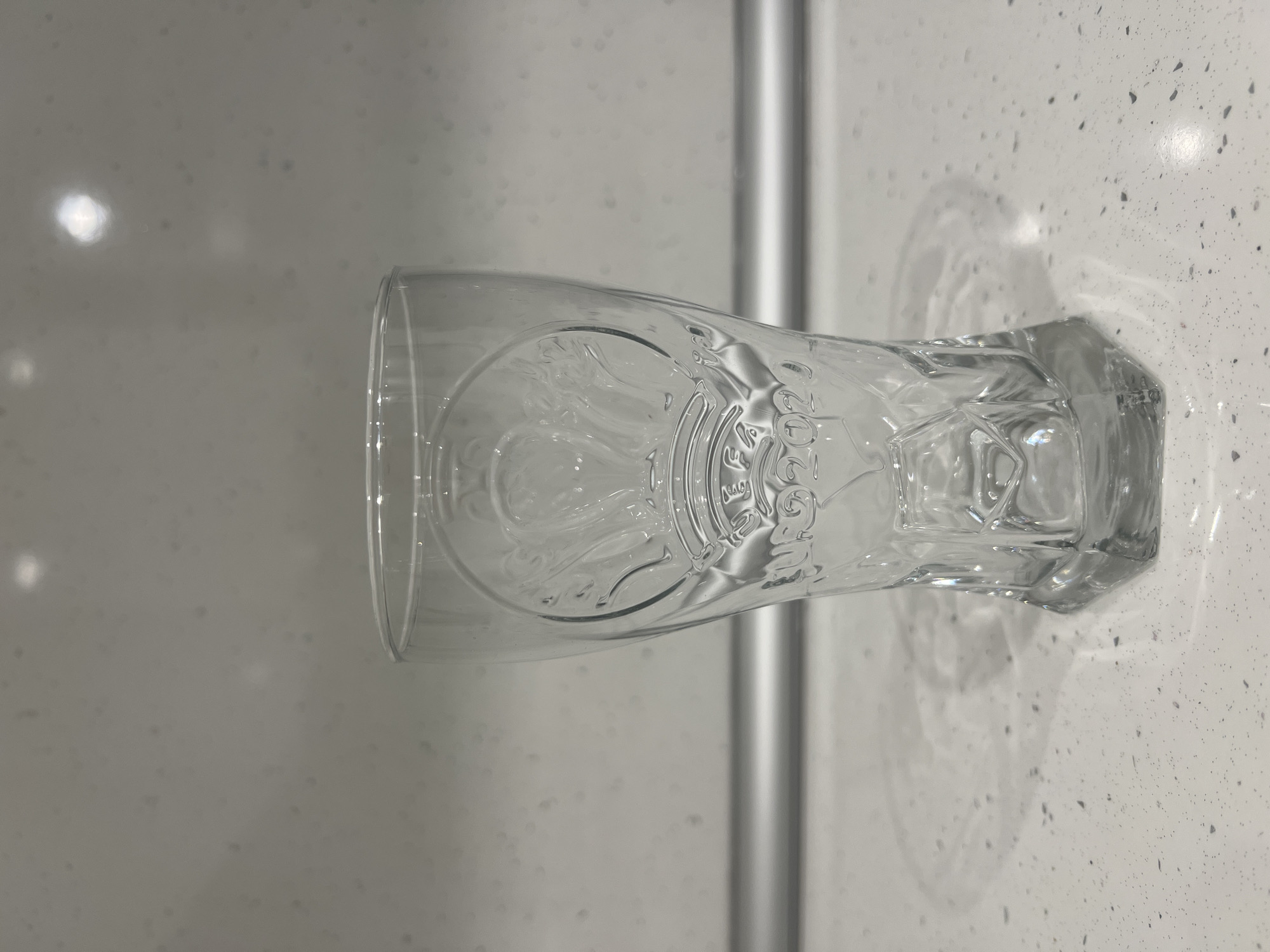 A clear glass tumbler with embossed detailing sits on a speckled white countertop. The glass features a design that includes what appears to be a crown or laurel wreath, and the text "UEFA 2020". Behind the glass, a silver bar runs horizontally across a light gray tiled wall. The countertop exhibits small dark speckles. The scene is illuminated by overhead lighting, casting a shadow of the glass onto the countertop. The location is a kitchen counter in Podgumer, Bulgaria. There are no people or actions depicted in the image.