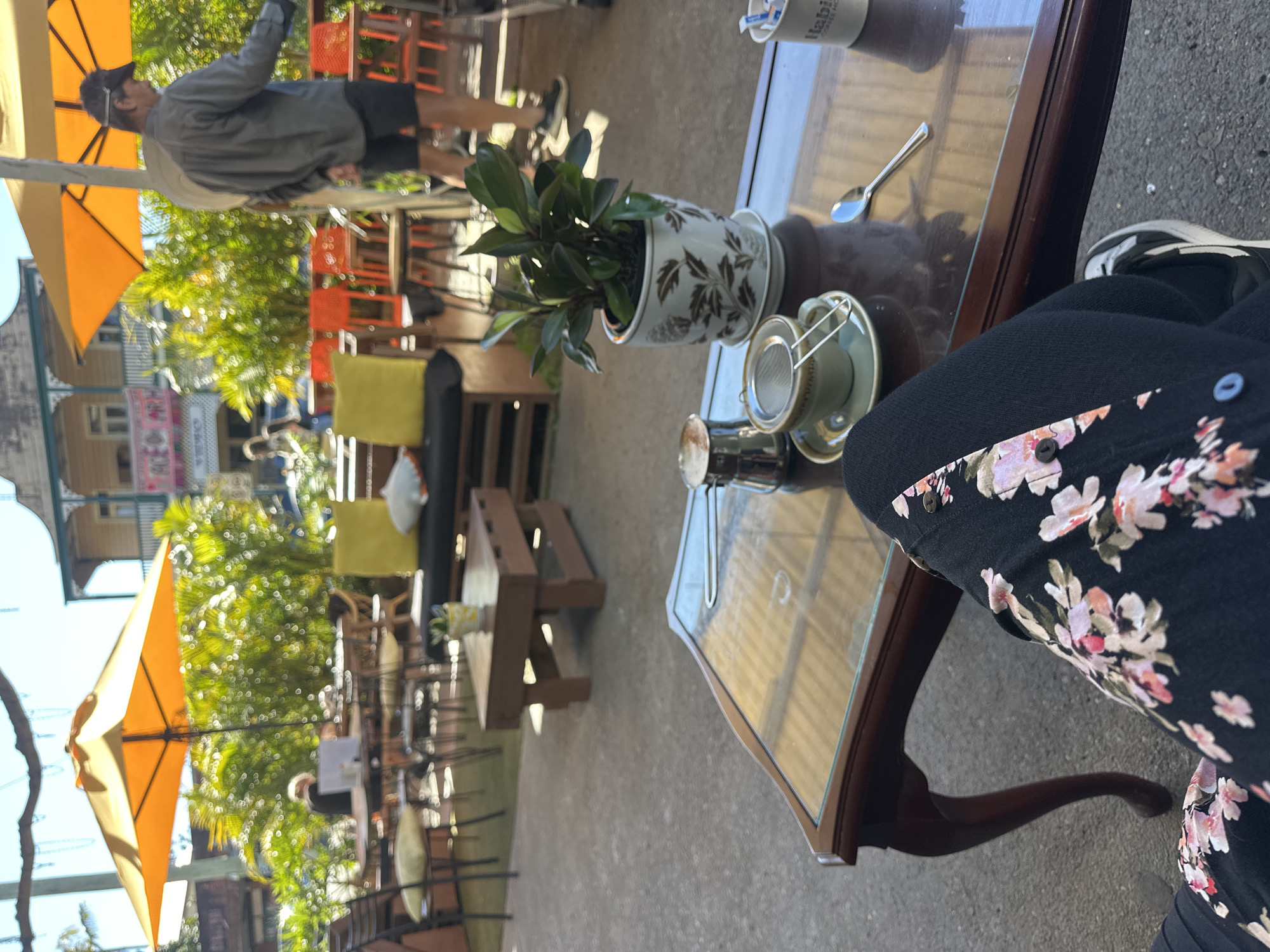 The image depicts an outdoor seating area at a cafe, likely in Bellingen, Australia, given the provided context. In the foreground, a person's legs are visible, wearing black pants with a floral pattern and sneakers. They are seated at a dark wood coffee table with a glass top. On the table, there is a teacup with a metal strainer, a small metal pitcher, a spoon, and a potted plant in a decorative white pot with brown floral designs. In the background, there are more tables and chairs, some with yellow cushions, under orange umbrellas. A building with a balcony and several windows is visible in the distance, along with lush green foliage. People are seen in the background, including a man in a gray shirt and shorts, and another person seated at a table. The overall atmosphere suggests a relaxed daytime setting at an outdoor cafe.