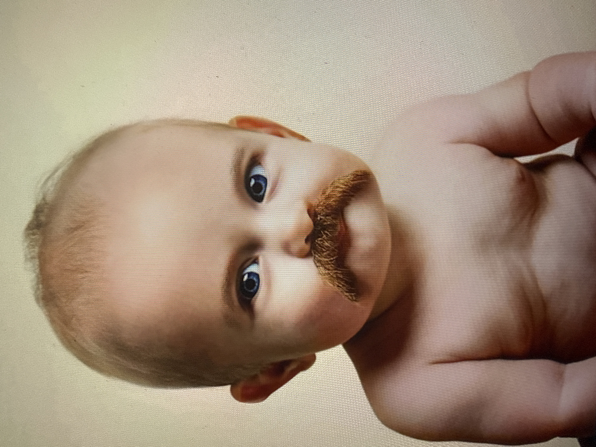 The image shows a close-up portrait of a baby wearing a fake mustache. The baby has fair skin, blue eyes, and is bald with a few wisps of blonde hair. The mustache is brown and bushy, extending across the upper lip. The baby is looking directly at the camera with a neutral expression. The background is a plain, light beige color. The image quality is slightly grainy, suggesting it may be a photograph of a screen or a lower-resolution digital image. The context of Tbilisi, Georgia, is not discernible from the visual information present in the image.