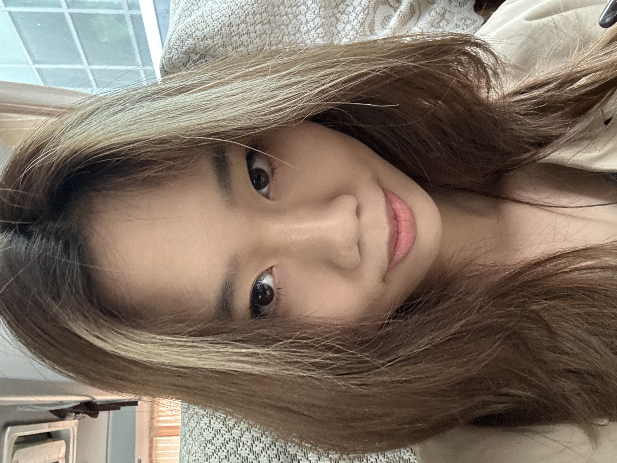 A close-up portrait of an Asian woman with shoulder-length brown hair with blonde highlights framing her face. She has brown eyes, fair skin, and slightly parted pink lips. The background includes a textured beige surface possibly a piece of furniture and part of a window with frosted or patterned glass. There is no information available in this image to indicate the location in Lanzhou, China.