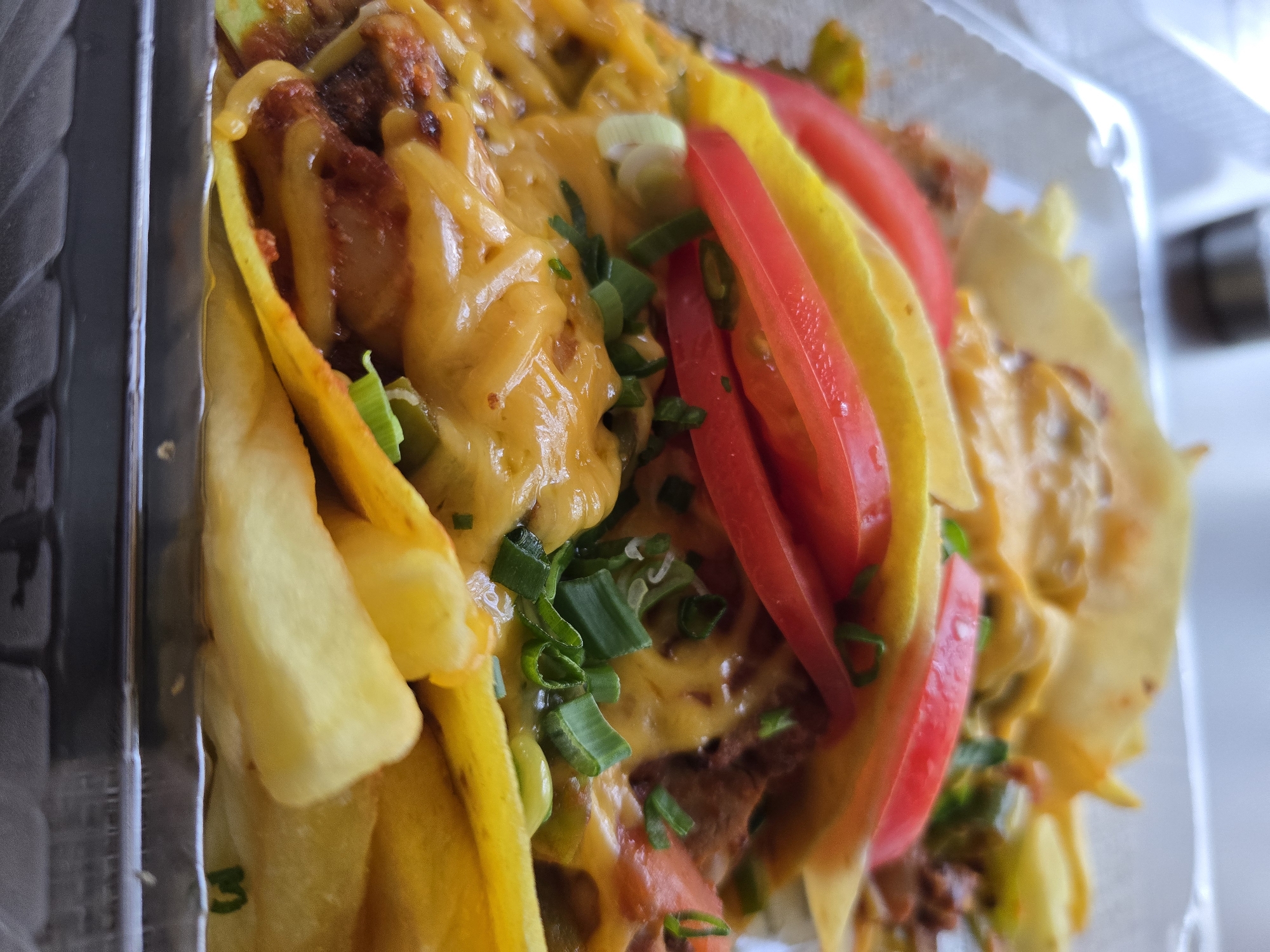The image shows a close-up view of food, likely a type of loaded fries or nachos, served in a clear plastic container. The main components are yellow fries, a meat-based topping (possibly beef or chicken), melted cheese, sliced tomatoes, and chopped green onions. The food appears to be freshly prepared. The location and additional context are unknown.