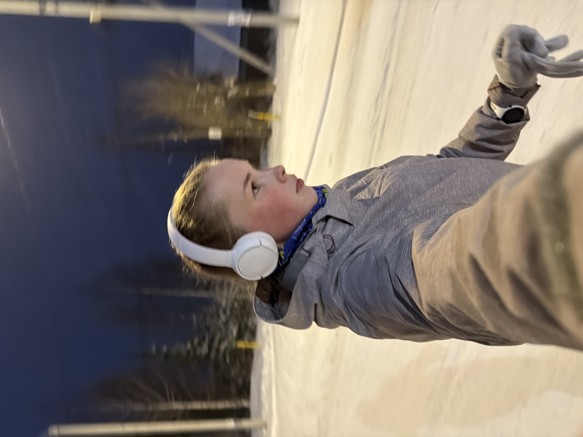 The image shows a young person wearing headphones, a gray jacket, and gloves. The person is positioned outdoors in a snowy environment. The background includes trees and an overcast sky transitioning from dark blue to a lighter gradient, suggesting either dusk or dawn. A horizontal white and gray structure is visible in the background. Given the location specification as Berezniki, Russia, the scene is likely a park or recreational area within the city during winter. The person appears to be taking a selfie.