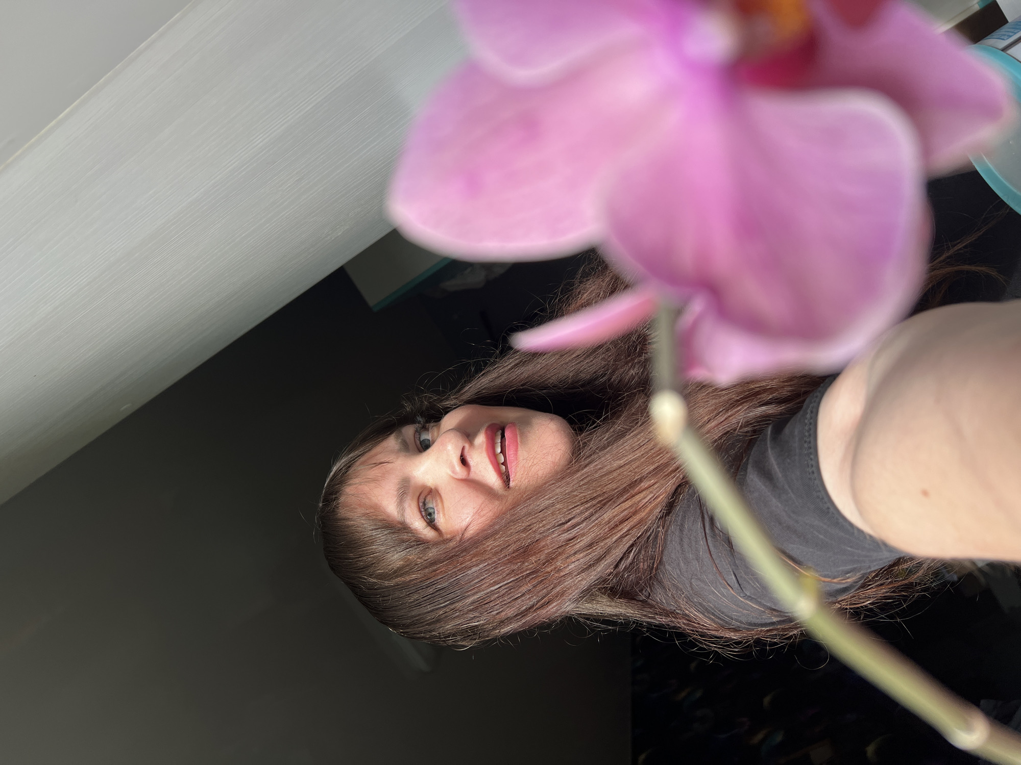 The image is a close-up portrait, oriented horizontally, featuring a woman and an orchid. The woman, with long, dark brown hair, is positioned on the left side of the frame, looking directly towards the viewer. Her blue or green eyes are open, and her lips are slightly parted, revealing a glimpse of her upper teeth. She has fair skin and is wearing a dark-colored, possibly black, short-sleeved top. Her expression is calm and neutral.
Dominating the foreground on the right is a large, blurry pink orchid flower, with a soft green stem extending downwards. The extreme blur of the orchid suggests a very shallow depth of field, indicating it is very close to the camera lens.
The setting appears to be indoors. The upper left background shows a light-colored, possibly white, surface with faint horizontal striations, which could be a wall or a piece of furniture. The remainder of the background is a dark, undefined area, creating a simple backdrop. There are no specific location cues visible within the image to directly indicate "Noviy, Russia." No text is visible in the media file. The lighting is soft and appears to be from an internal source, enhancing the focus on the subjects.