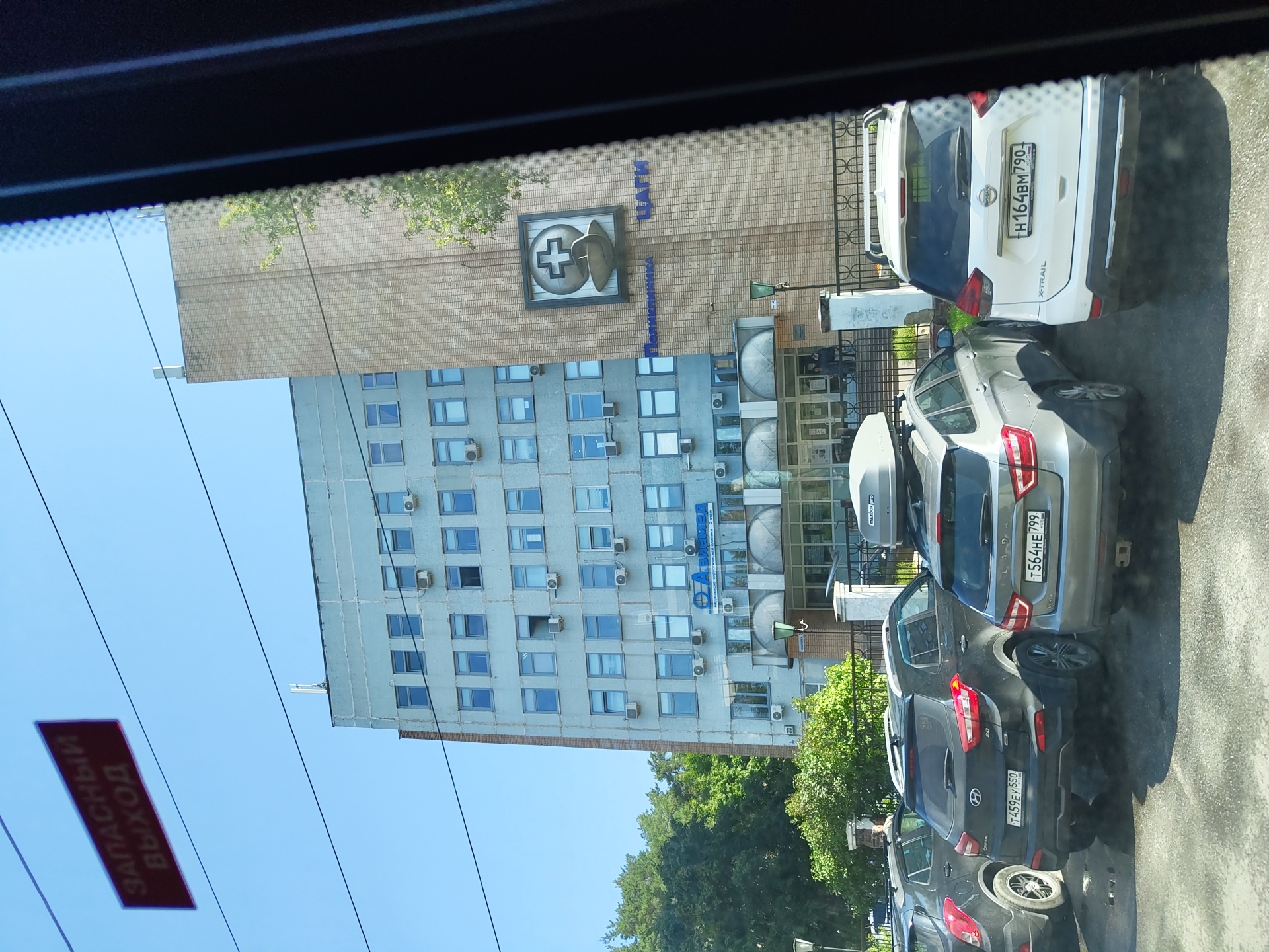 The image shows a view of the exterior of a medical facility, likely a clinic or hospital, in Zhukovsky, Russia. The building is a multi-story, light-blue concrete structure with numerous windows and visible air conditioning units. On the facade, there is a blue sign with Russian text, the larger part reading "АЛИМЕД" and a smaller line below it "МЕДИЦИНСКИЙ ЛЕЧЕБНО-ДИАГНОСТИЧЕСКИЙ ЦЕНТР 2 ЭТАЖ." To the right, on a brick-clad section of the building, a logo depicting a medical cross above a stylized bowl is visible, with the Russian words "Поликлиника ЦАГИ" below it. Several cars are parked in front of the building, including a black Hyundai Creta, a silver Lada Vesta, and a white Nissan X-Trail. A red sign with white text reading "ЗАПАСНЫЙ ВЫХОД" (Emergency Exit) is suspended in the sky, likely attached to overhead wires. The sky is clear and blue, and green trees are visible behind the building. The image is taken from inside a vehicle, suggested by the tinted window frame on the right side of the frame and the reflections on the glass.