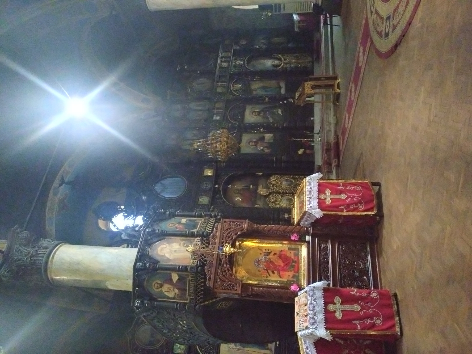 The image shows the interior of an Orthodox church, possibly in Stara Zagora, Bulgaria, given the context. The scene is dominated by a large iconostasis adorned with numerous religious icons, with a chandelier hanging in front. Two stands covered in red cloth with gold crosses are positioned in the foreground. A marble column is visible on the left. The floor is made of wood, and a rug is present in the background. The interior is dimly lit, with a bright light source near the ceiling.