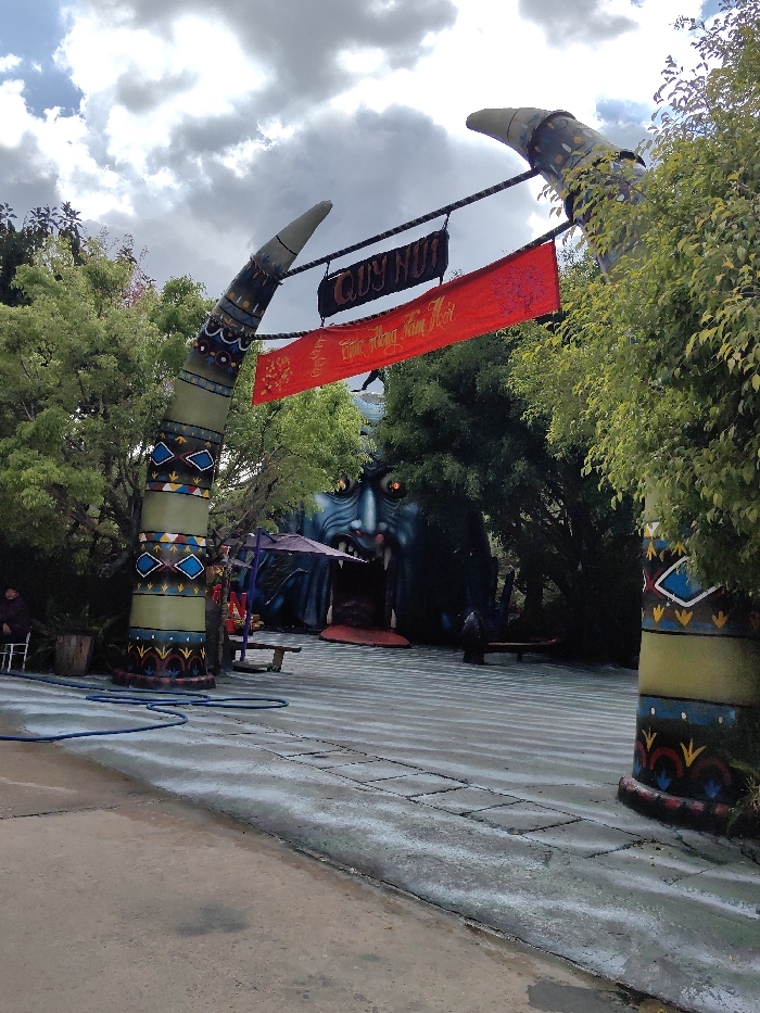 The image depicts an outdoor entrance area in Păng Tiêng 1, Vietnam, under an overcast sky. A large archway, resembling two curved tusks or horns, stands prominently in the foreground. These structures are decorated with multicolored geometric patterns in shades of green, brown, blue, yellow, and red. Hanging from the archway are two banners: a black banner with white text reading "QUY NUI" and a red banner beneath it with yellow/gold text, likely Vietnamese. Beyond the archway, a dark blue/grey structure designed as a monstrous face with an open mouth serving as an entrance and glowing red eyes is partially obscured by dense green foliage. A dark purple umbrella is visible to the left of the monster-face entrance. The ground in the foreground consists of a concrete path, which transitions to a striped paved area leading toward the monster-face structure. A coiled blue hose is present on the concrete path on the left. One individual, dressed in dark clothing, is seated on a white plastic chair on the far left, positioned next to a wooden barrel. A dark, indistinct figure is partially visible near the base of the monster-face entrance on its right side.