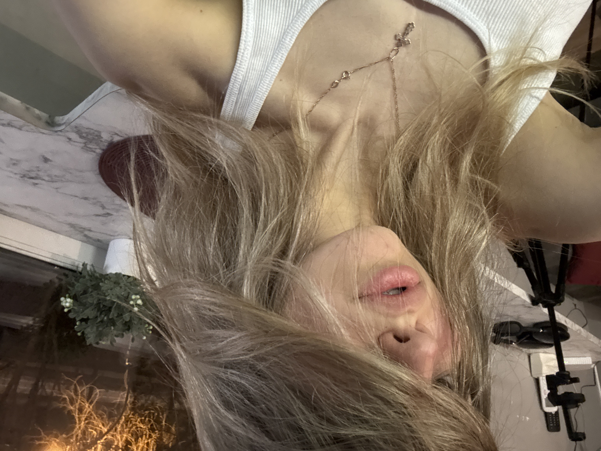 A close-up, inverted image shows a woman with long blonde hair wearing a white tank top and a delicate gold necklace with a small cross pendant. The background includes a window with foliage visible outside, a marble-patterned countertop, and miscellaneous objects including what appears to be a tripod. There is no information in the image that suggests a location in the city of Khimki, Russia.