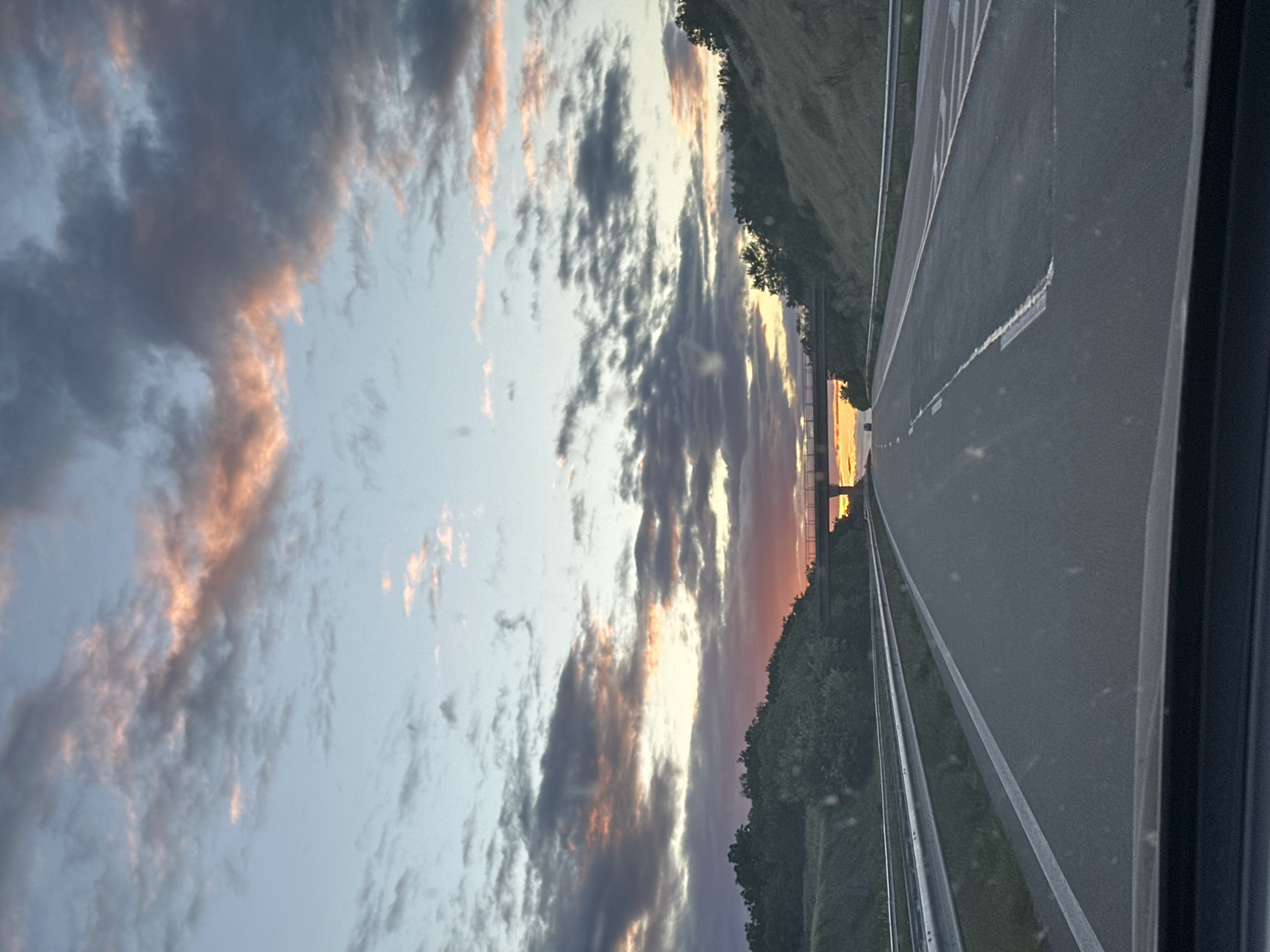 The image shows a view from a vehicle traveling on a highway, possibly near Dobrich, Bulgaria. The road is asphalt, with visible lane markings. Along the roadside, there's vegetation, possibly trees and bushes, marking the edge of the road. Above, the sky is filled with clouds, illuminated by the colors of a sunrise or sunset, displaying hues of orange and gray. The image appears to be taken through a window, as evidenced by a dark frame on one side.