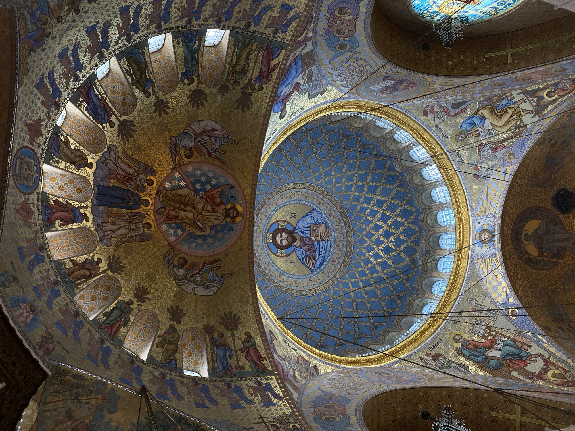 The image shows the interior of the Naval Cathedral of Saint Nicholas in Kronstadt, Russia, looking up at its domes and ornate decorations. The main focus is on the large central dome, which features a mosaic image of Jesus Christ in blue and gold. Surrounding this dome, and on the smaller domes and arched ceilings, are numerous mosaics depicting religious figures, angels, and intricate patterns in gold, blue, red, and other vibrant colors. Sunlight streams in through arched windows along the upper edges of the domes, illuminating the detailed artwork. Several chandeliers hang from the ceiling. The overall impression is one of grandeur and spiritual richness, characterized by the extensive use of mosaics and gold leaf.