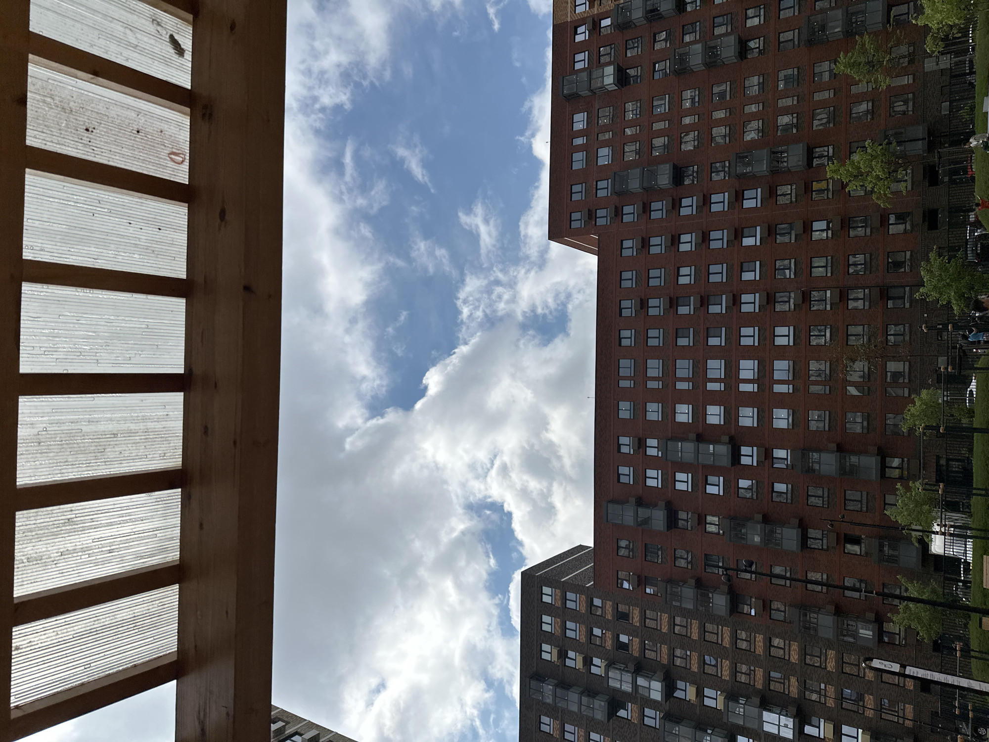 The image depicts a view from beneath a wooden pergola with a translucent, corrugated roof, looking up at a partly cloudy sky and two large apartment buildings constructed of brick. The building on the left is darker brick, and the building on the right is reddish-brown brick. Both buildings have numerous windows and balconies. The scene appears to be in a residential area. There are trees and a black metal fence in the foreground, with some people and vehicles visible beyond the fence. The image quality is clear.