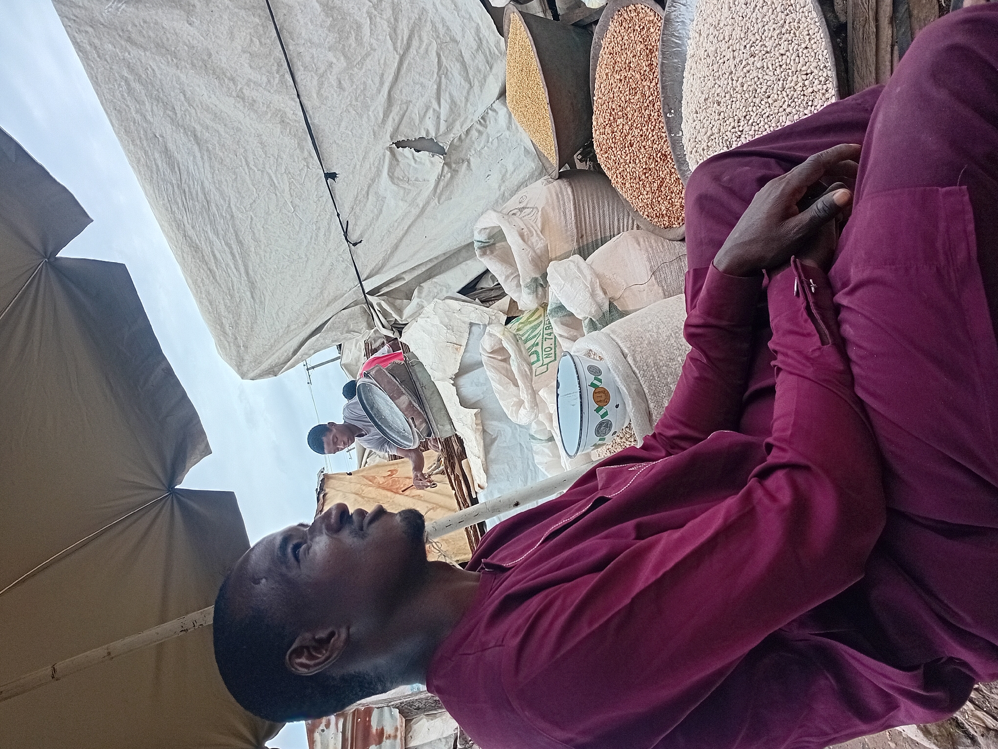 An outdoor market scene in Maiburiji, Nigeria, features a man in a burgundy traditional outfit seated under a large tan umbrella. He is facing to the left in profile, with his hands resting on his lap. In the background, another man is visible, tending to a sieve containing grains. Several large sacks filled with assorted grains are arranged around the foreground and middle ground, along with large circular metal trays overflowing with grains. The market appears to be set up with temporary structures, including a large white tarp covering a portion of the background. The sky is overcast.