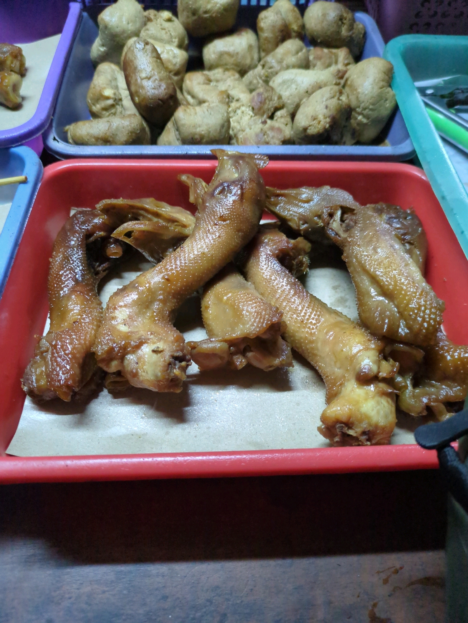 This is a high-angle, eye-level, medium shot of several trays of food, likely at a street food stall in Sindon, Indonesia. The foreground features a red plastic tray filled with cooked duck feet, which are glistening and appear to be glazed or sauced. The duck feet are arranged in a cluster, with some overlapping. In the background, to the left, is a purple plastic tray with a pile of brown, irregularly shaped food items, possibly fried tofu or some type of fritter. To the right of the purple tray, and slightly behind it, is a teal-colored plastic tray with some green utensils visible. The overall scene suggests a food vendor or market stall environment. There are no people visible in the image.