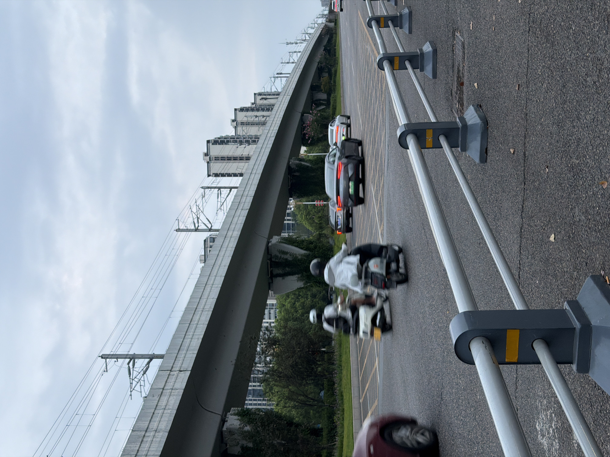 The image depicts a street scene in Jiangjin, China, under an overcast sky. A prominent elevated structure, likely a viaduct for a railway or highway, runs diagonally across the frame from left to right, supported by concrete pillars overgrown with vegetation. Overhead power lines associated with the elevated structure are visible. On the road below, several vehicles are in motion. Two individuals are riding scooters, one white and one dark gray, in the foreground lanes. Behind them, a dark gray car with illuminated red taillights is moving forward. Further down the road, a white car and another dark vehicle are also visible. The road is paved with asphalt and marked with yellow lines. A grey metal barrier with yellow reflective markers runs along the right side of the road in the foreground. Tall buildings, characteristic of an urban environment, are visible in the background beyond the elevated structure and trees lining the street. The overall atmosphere is one of a typical urban commute.