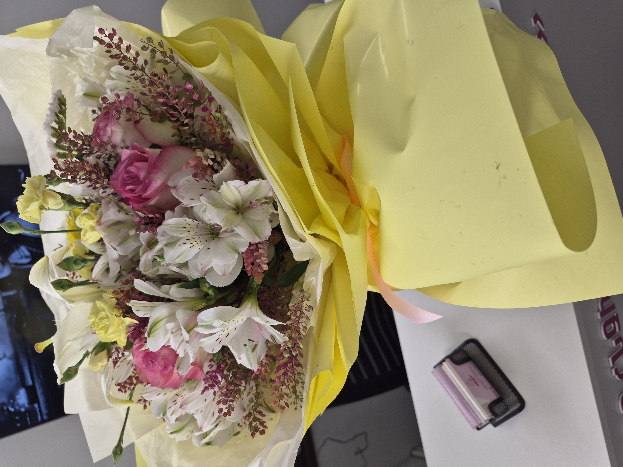 The image captures a vibrant bouquet of mixed flowers, resting on a light-colored surface, likely a desk or table, in an indoor setting in Yerevan, Armenia. The bouquet is prominently wrapped in bright yellow paper, secured with a delicate pink ribbon, and features an inner layer of white tissue paper.
The floral arrangement includes several distinct types of flowers: two prominent pink roses with lighter edges, numerous white alstroemeria (Peruvian lilies) characterized by their distinctive speckled patterns, and smaller clusters of yellow flowers, possibly carnations or chrysanthemums. A dark reddish-purple, fine-leaved foliage, acting as a filler, adds depth and contrast to the bouquet.
In the background to the right, a black mesh organizer holds a stack of small, light pink or purple cards, which appear to be business cards. Further right, a partial, vertical sign or object displays silver-colored text against a gray background, clearly showing the numbers "46". To the left of the bouquet, a blurry, dark blue and black reflective surface is visible, possibly a monitor screen or a window.
The scene is brightly and evenly lit, suggesting daytime. No specific activity is taking place; the image is a static portrayal of the bouquet and its immediate surroundings.