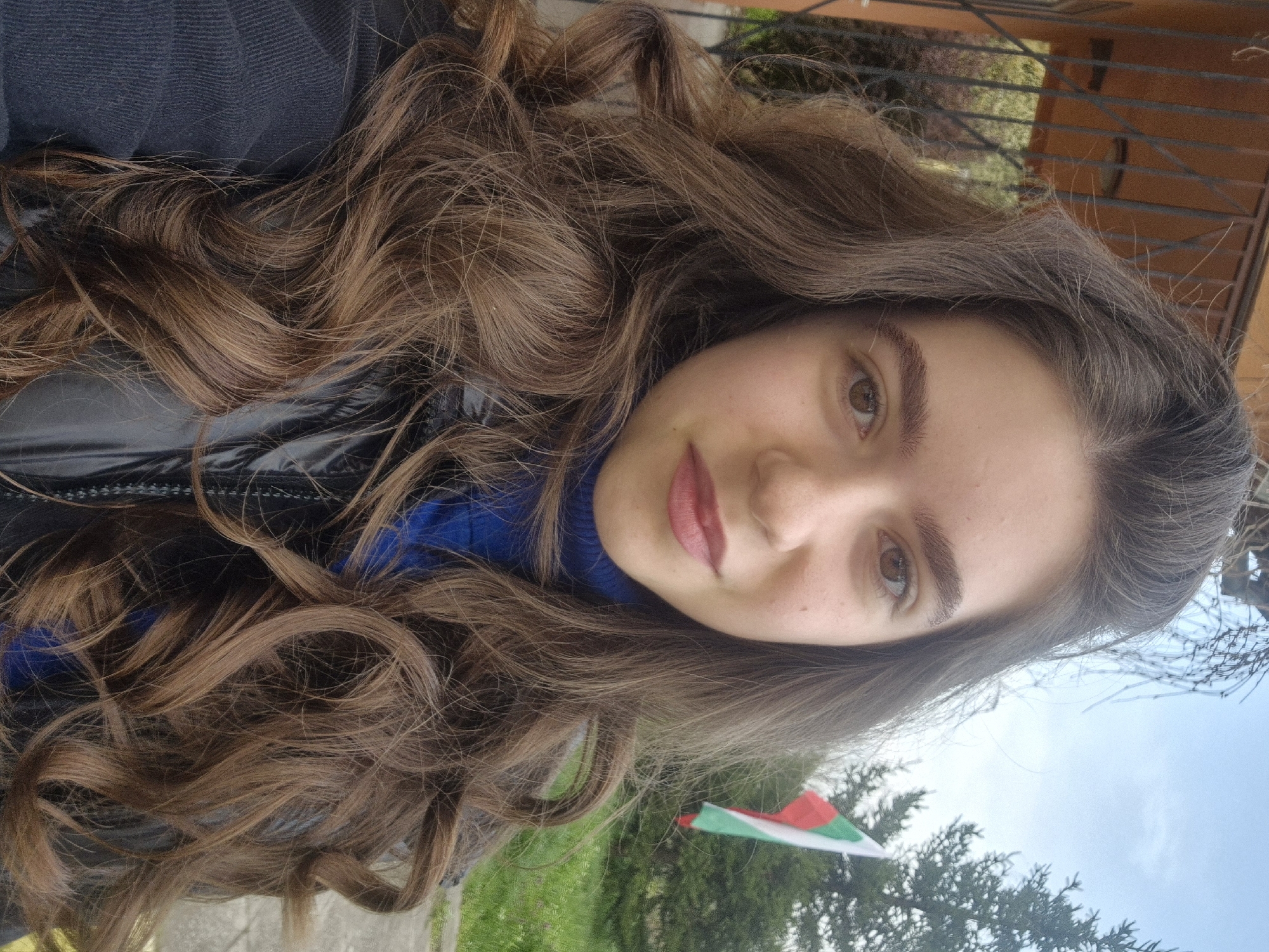 A young woman poses for a selfie outdoors. She has long, wavy brown hair and is wearing a blue top under a black, shiny jacket. In the background, there is greenery, a fence, and a structure possibly belonging to a house. A Bulgarian flag is visible. It can be inferred from this flag that the location is in Bulgaria, specifically Pavlikeni.