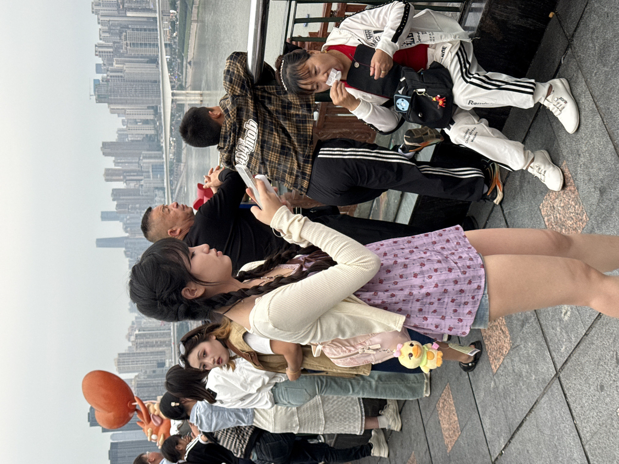 The image depicts a group of people standing along a waterfront in Yuzhong District, China, with a cityscape in the background. From the front, a woman with braided hair looks to her left, holding a smartphone. Behind her are several other individuals, including a woman in a white top and vest with sunglasses, a person in a white shirt and blue jeans, and a person in a white dress. Further back, there's a person holding a heart-shaped balloon and an orange character toy. In the foreground, a woman in a white jacket and black and white striped pants is eating something. Next to her, a person in a checkered jacket leans against a railing. The setting appears to be a public space by the water, possibly a promenade or viewing platform. The weather seems overcast.