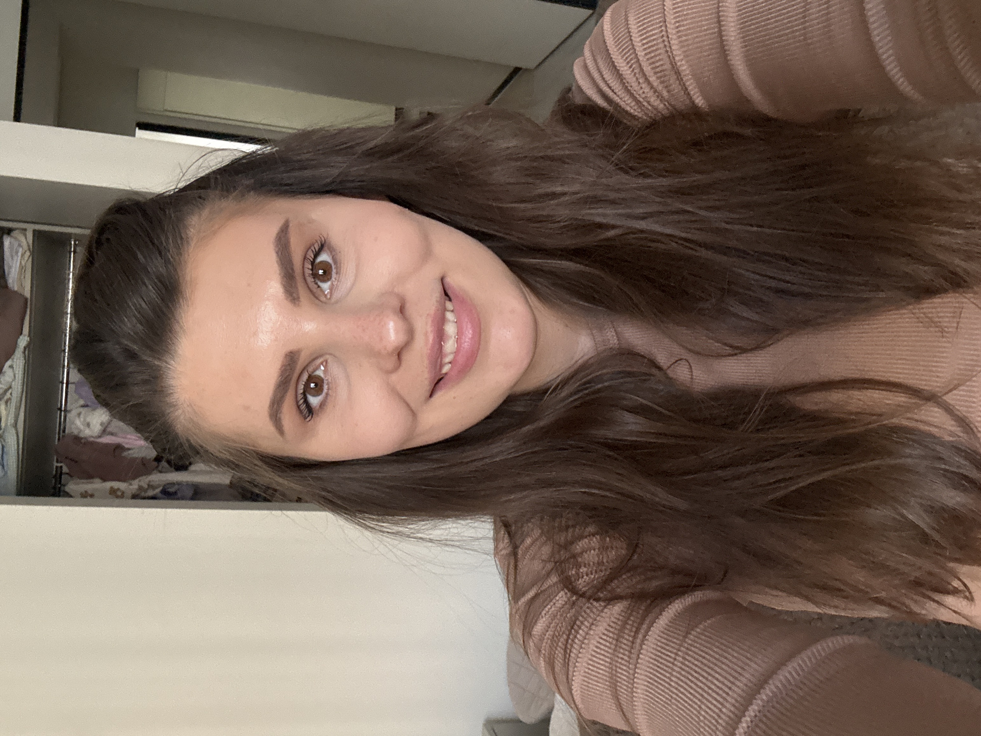 A medium close-up selfie captures a young woman with a friendly, broad smile, looking directly at the camera. Her long, dark brown hair, styled with a slight wave, cascades over her shoulders and chest. She has expressive brown eyes, framed by well-defined eyebrows and visible eyelashes enhanced with mascara. Her complexion is fair, and she sports a subtle blush. She is wearing a light peach or blush-toned ribbed knit top, the distinct texture of which is visible on her garment.
The setting is indoors, with a softly lit, somewhat blurred background. Behind her left shoulder, a glimpse of an open closet or shelving unit reveals neatly arranged clothing, suggesting a bedroom or personal living space. The remainder of the background consists of plain light-colored walls or furniture. The lighting is even and diffused, typical of indoor conditions, with no specific indicators of the time of day. The woman's demeanor is cheerful and approachable. There is no visible text within the image. This scene is located in Sofiivska Borshchahivka, Ukraine.