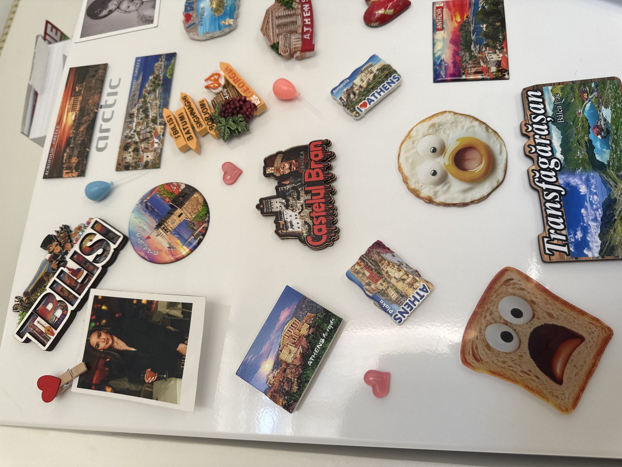 A white refrigerator door is covered with various magnets. In the upper left quadrant, a framed photo shows a woman holding a drink. To its right, a magnet depicts the word "TBILISI" in a stylized font. Below the photo is a round magnet showing a cityscape and a historical monument. In the upper center, a magnet displays "Acropolis, Greece," and below it, another magnet shows a scenic view of a Greek village. Further down, a directional sign magnet points to "Tbilisi," "Batumi," "Sighnaghi," and "Borjomi." To the right of the "Tbilisi" magnet, a magnet with a baby's face is visible. Several smaller magnets are scattered across the door, including a red heart, a blue balloon, a pink balloon, a magnet with a fried egg, and a magnet with a slice of toast with a shocked expression. Several other magnets depict architectural landmarks and landscapes, including "Castelul Bran," "Athens Acropolis," "Plaka Athens," "Athens," "Cyprus," and "Antalya." A large magnet at the bottom features a landscape with a lake and text that reads "Transfăgărășan Bâlea Lac." The scene is a close-up of a refrigerator door adorned with travel souvenirs. No information is provided to indicate the location is Chișinău, Moldova.