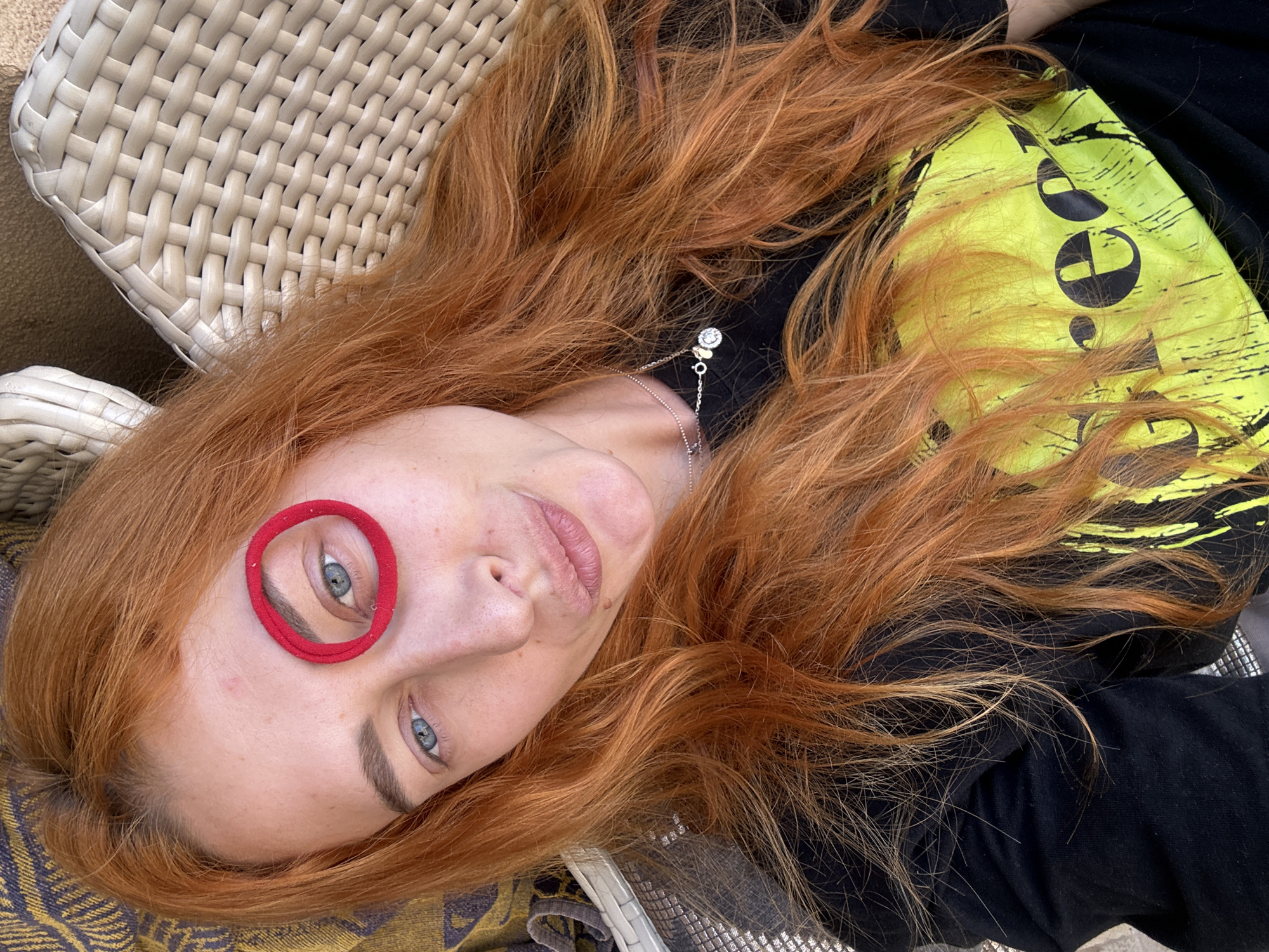 A woman with long red hair is lying down, angled slightly upwards, on a light-colored wicker surface. She has a red hair tie around her left eye. She is wearing a black shirt with a yellow design. A silver necklace is visible around her neck. The image appears to be taken outdoors during the daytime. The location in the city of Budva, Montenegro cannot be determined.