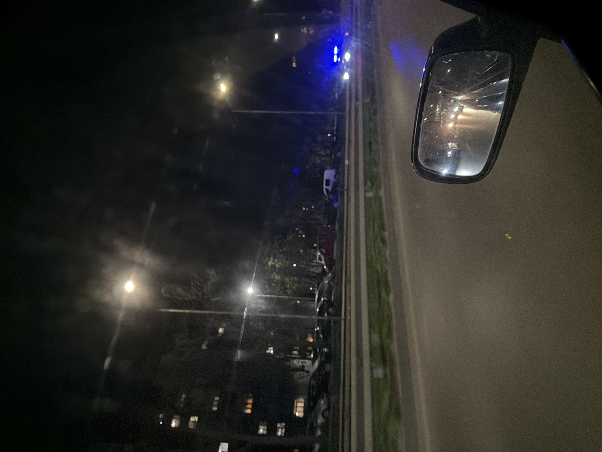 The image, taken from inside a vehicle at night, shows a road scene. Streetlights illuminate the road and surrounding buildings. A rearview mirror on the right side of the frame reflects lights from other vehicles. In the distance, there are blue lights that are likely from an emergency vehicle. Along the left side of the road, a building with lit windows is visible. Cars are parked along the side of the road. The image was taken in Varna, Bulgaria.