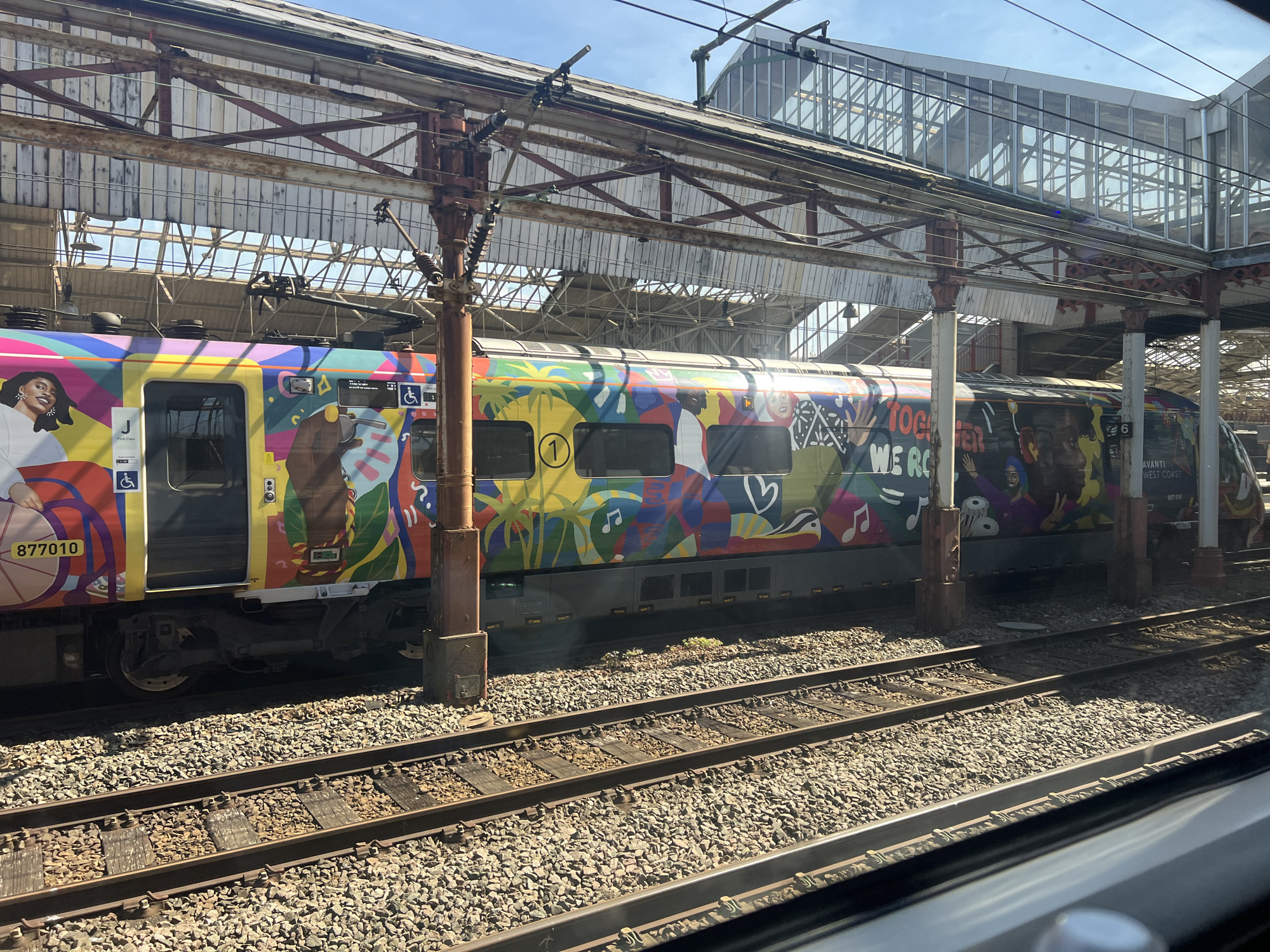 The image shows an Avanti West Coast train at Crewe railway station in Crewe, Cheshire, UK. The train has a colorful design with figures and images. Tracks run in the foreground. Above the train is the steel framework of the station roof and a footbridge. The sky is blue and sunny.