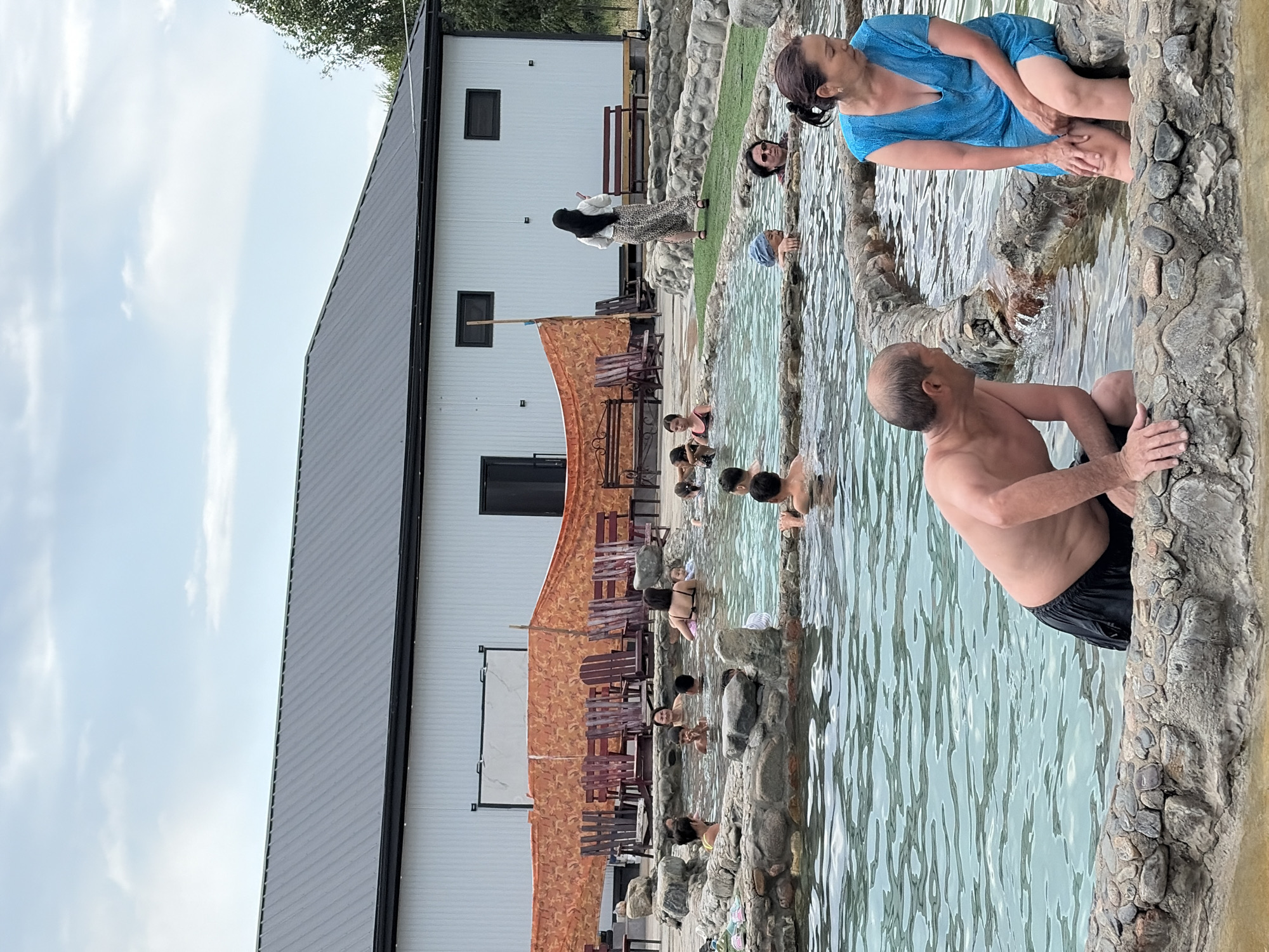 An outdoor hot spring facility features a man and a woman sitting on the rocky edge of a pool. The man, bare-chested and wearing black shorts, is partially submerged in the water and looking towards the woman. The woman, wearing a blue dress, is seated on the edge, with her hands resting on her knees. Several other people are visible in the pool behind them, enjoying the water. The pool is surrounded by a decorative rock border, and a white building with a dark roof is visible in the background. Orange netting and wooden chairs are set up in front of the building. The sky above is partly cloudy.