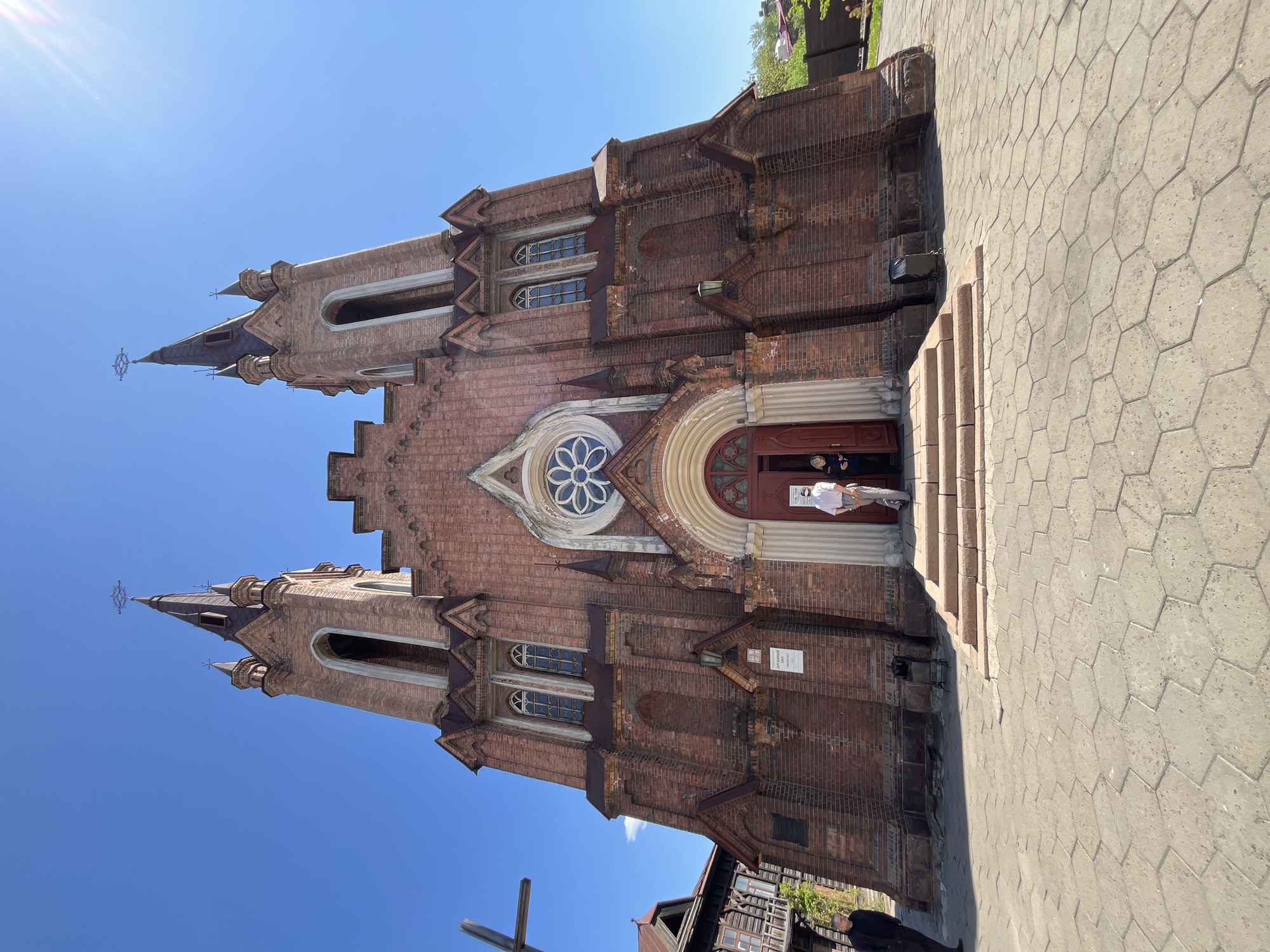 The image shows the Roman Catholic Church of the Transfiguration in Krasnoyarsk, Russia, viewed from a low angle, slightly off-center. The church is made of red brick, with two towers topped with spires and crosses. A person stands at the entrance. The foreground includes a hexagonal-tiled pavement and a corner of a building on the lower left. The sky is blue.