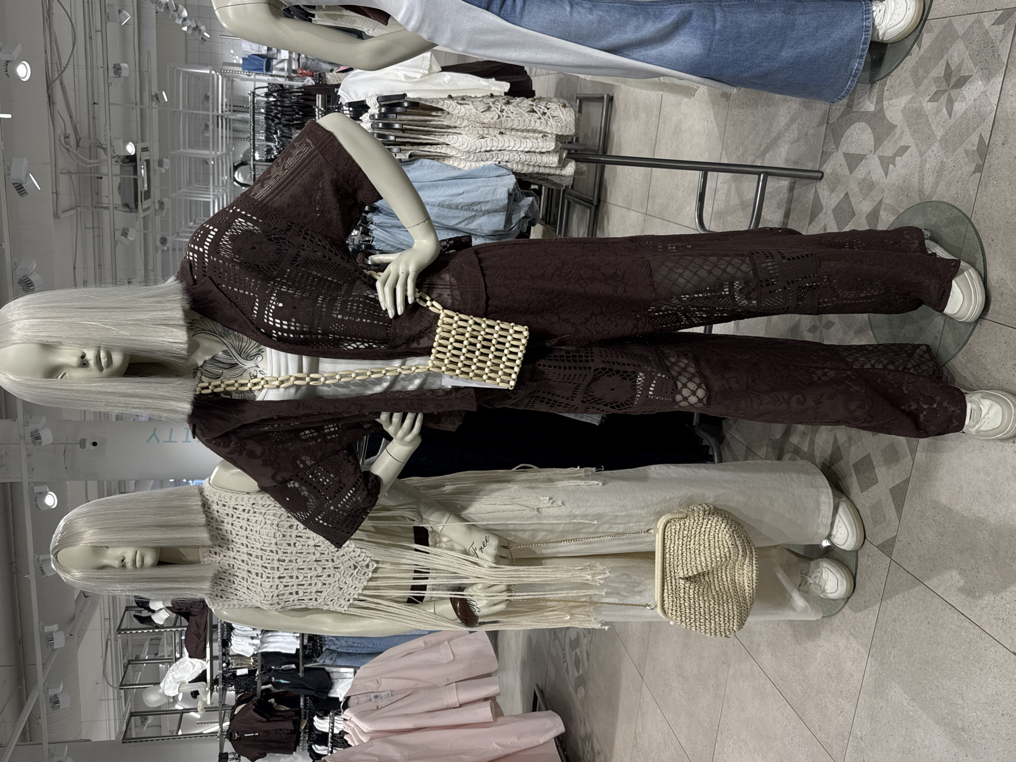 The image depicts two mannequins in a store setting, likely a clothing retailer. The location appears to be in Vsevolozhsk, Russia, based on potential product offerings and store design.
The upper mannequin is dressed in a brown lace-like outfit, including a top or jacket and pants, accessorized with a small, light-colored beaded bag. The lower mannequin is wearing a white, crocheted-style top with fringe, white pants, and carrying a woven straw-like bag. The mannequins have simple, light-colored wigs.
In the background, there are racks of clothing displayed for sale. The store has bright, industrial-style lighting. The flooring is a neutral tile with a geometric pattern.