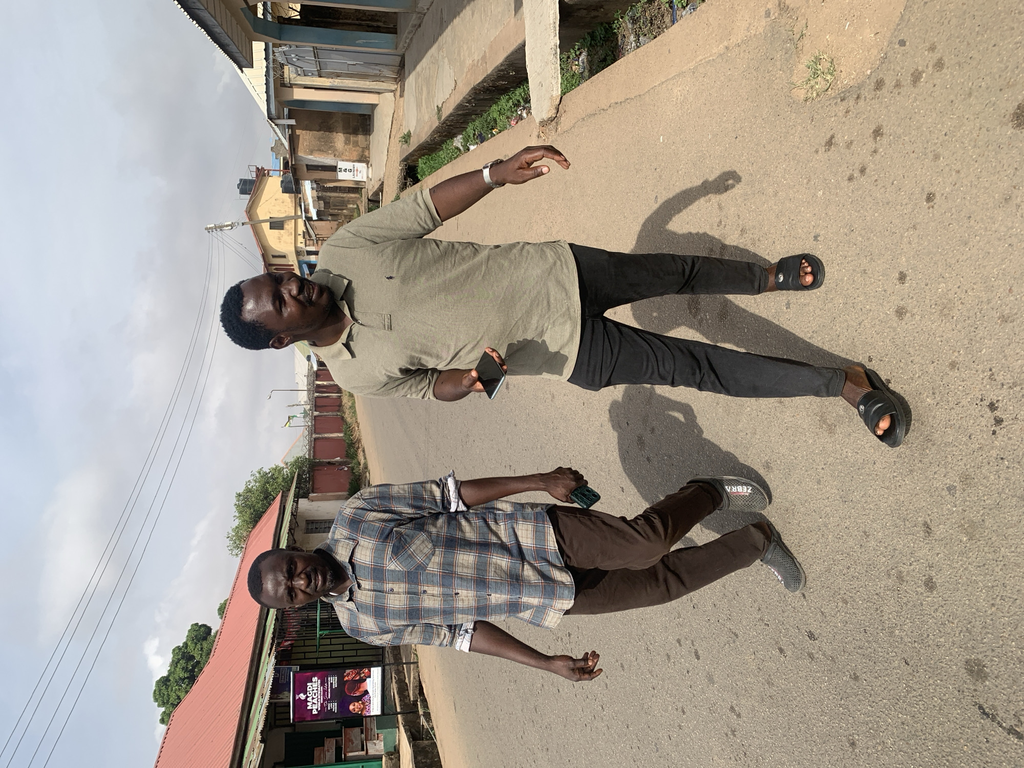 This image, taken in Jos, Nigeria, depicts two men standing on an asphalt road under an overcast sky, in what appears to be an urban or semi-urban environment. The photo is rotated 90 degrees counter-clockwise from its natural upright orientation.
The man in the foreground (right when upright) wears an olive green polo shirt, black trousers, and black slide sandals. He has short dark hair, a beard, and holds a black smartphone in his left hand, while wearing a silver watch on his left wrist. His right hand is slightly raised in a relaxed gesture.
The second man, positioned behind and to the left of the first, wears a blue and white plaid long-sleeved shirt with rolled-up sleeves, brown trousers, and dark grey sneakers that have "ZEBRA" written on the side of one shoe. He also has short dark hair and a beard, and holds a green-backed smartphone in his right hand. Both men cast distinct shadows on the road, indicating the sun's position. They appear relaxed, with neutral to slightly smiling expressions.
The background shows various buildings, many with corrugated metal roofs and concrete walls, common in the region. A ditch or gutter containing some greenery and debris runs along the right side of the road. Utility poles and wires crisscross the hazy sky.
Notable visible text includes a poster behind the second man, advertising "MAGDI PEACHES" with images of individuals and smaller accompanying text. A smaller, partially obscured sign in the distance also appears to display "M & G". The scene captures a casual moment on a street.