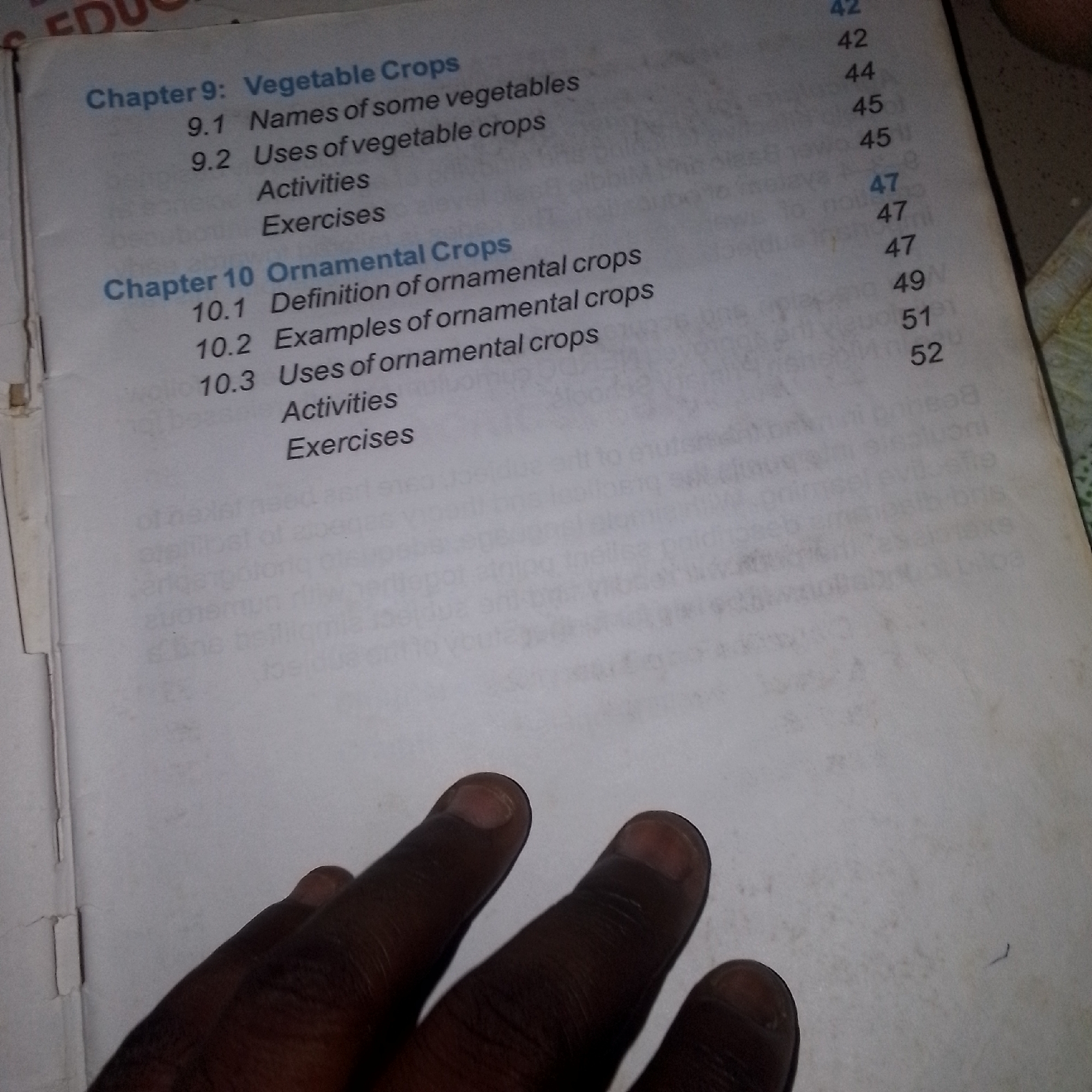 The image displays a page from a book, likely a textbook, with a table of contents or index. The page is partially obscured by a hand with dark skin and visible fingernails, which is positioned in the lower left portion of the frame. The book appears to be open to pages with chapter titles and subheadings, including "Chapter 9: Vegetable Crops" and "Chapter 10: Ornamental Crops." Corresponding page numbers are listed to the right of the subheadings. The environment is not discernible beyond the context of a book and a hand. The image is taken from a close-up perspective, focusing on the book's content and the hand.