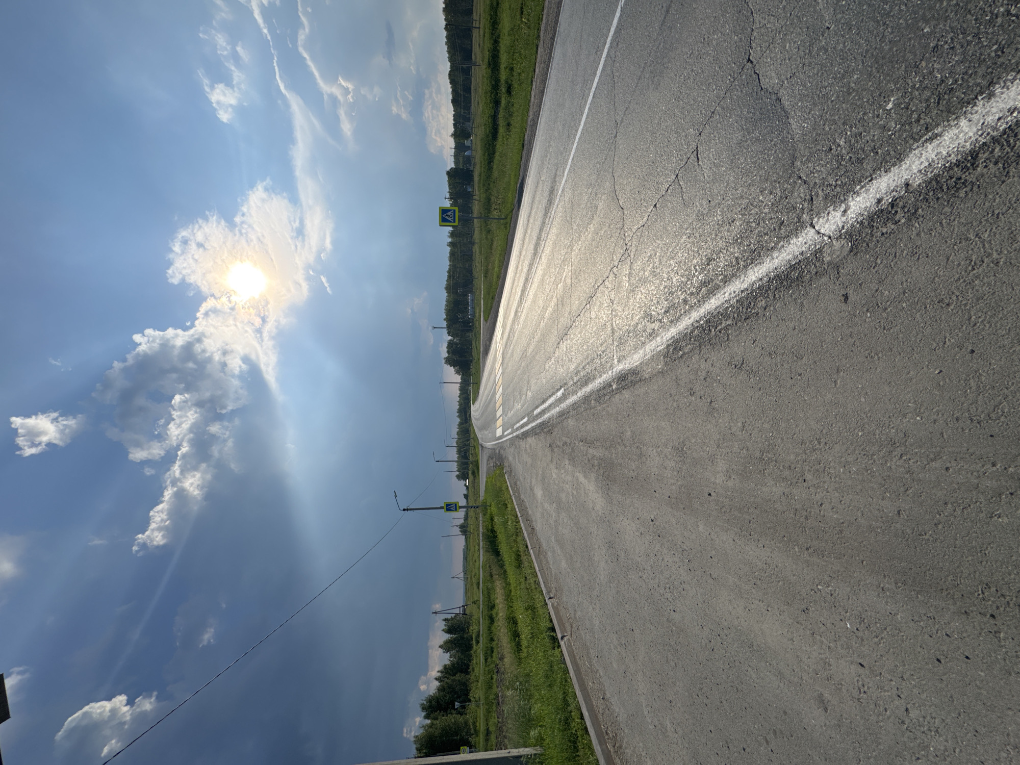 The image depicts a rural scene on a sunny day, likely in Arkhangelskoye, Russia. The perspective is from ground level, looking down a paved road that curves gently to the right and into the distance. The road is marked with a white line on the right edge and appears to be wet in some sections, reflecting the bright sunlight. The sun is high in the sky, casting strong rays and creating lens flare, indicating it's likely midday. Fluffy white clouds are scattered across a vibrant blue sky.
To the left of the road is a strip of lush green grass, followed by a line of trees that create a distant horizon. A pedestrian crossing sign, a yellow diamond with a black silhouette of a person walking, is visible on a pole alongside the road. Other poles, possibly for streetlights or power lines, are also scattered in the background. The overall mood is peaceful and serene, with the bright sunshine and natural landscape creating a pleasant atmosphere. No people are visible in the frame, and the focus is on the road and its surrounding environment. There is no visible text in the image.