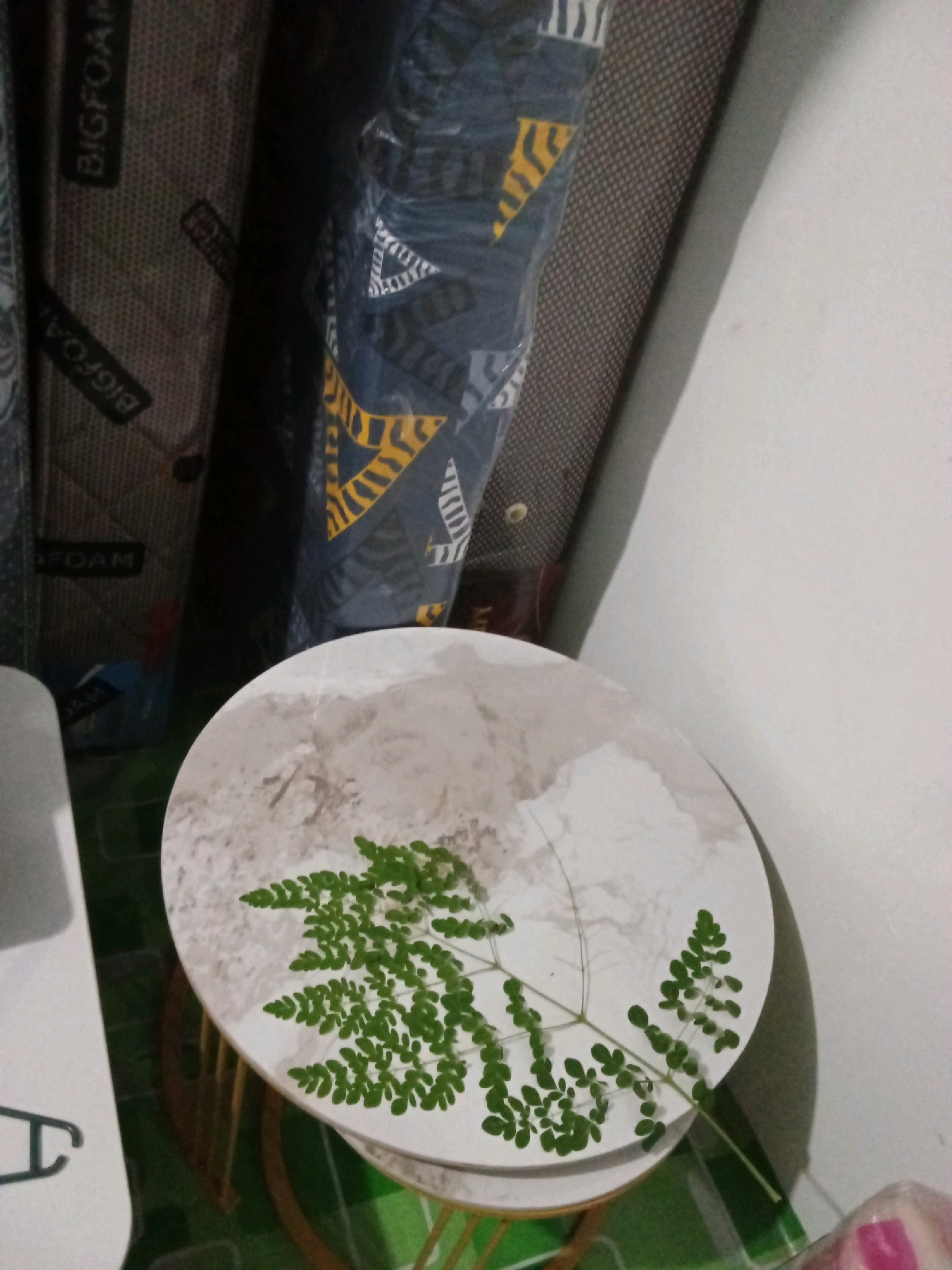 The image shows a round side table with a marble-like pattern, topped with a sprig of green leaves. The table is placed against a white wall, with several rolled-up mattresses visible in the background. Some of the mattresses have patterned fabric, with text that appears to be "BIGFOAM" visible on at least one. The flooring is a green and dark patterned surface. The lighting suggests an indoor environment. There are no people visible in the image. The scene does not provide enough information to determine the specific location within Gas Alam Badak I, Indonesia.