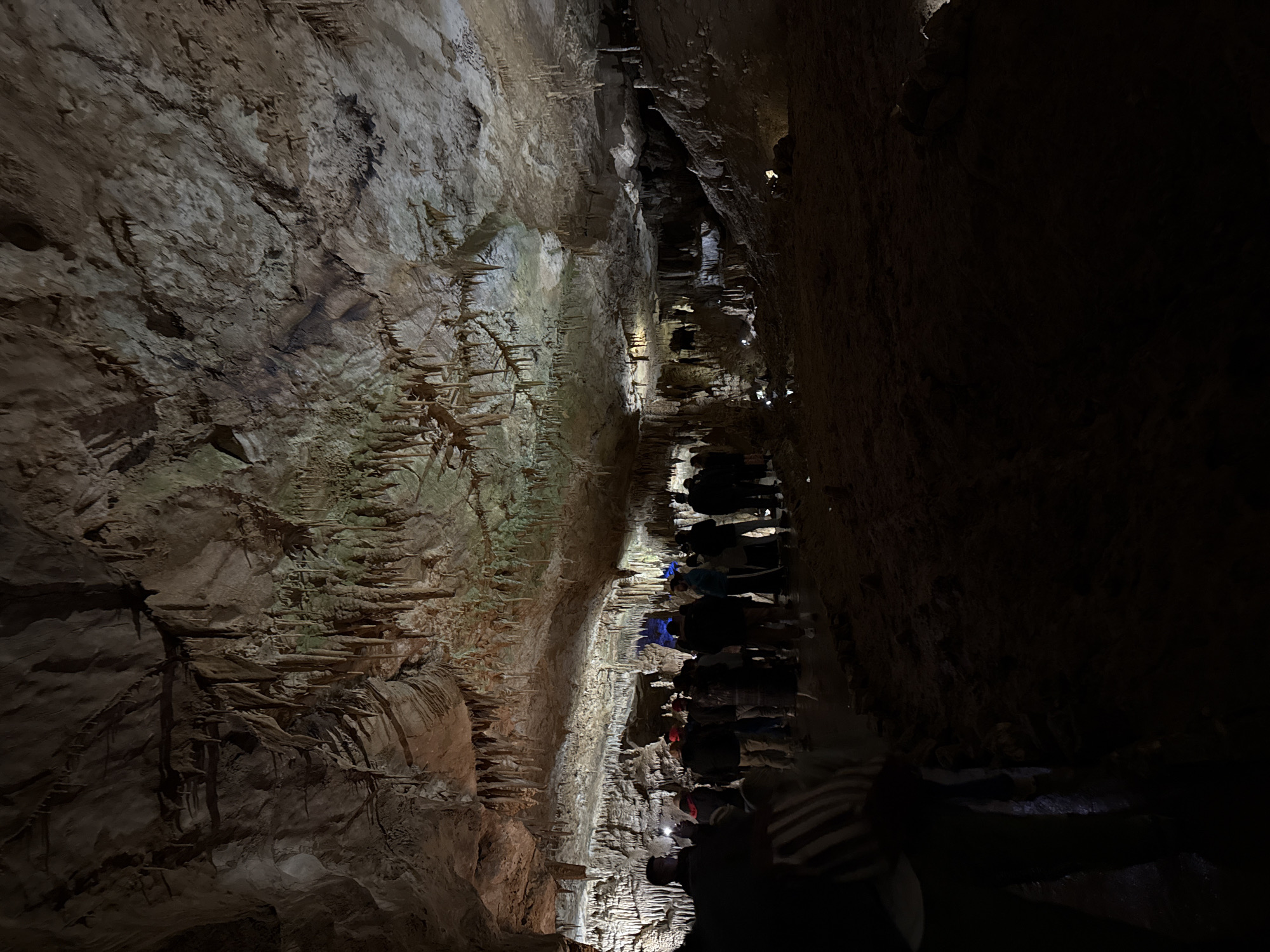 A group of people is walking through the Prometheus Cave in Tskaltubo, Georgia. The cave walls are rough and covered in stalactites and stalagmites. The lighting is dim, with some areas brightly lit and others in near darkness. People are wearing a variety of clothes. The walkway seems narrow, with cave formations very close on both sides.