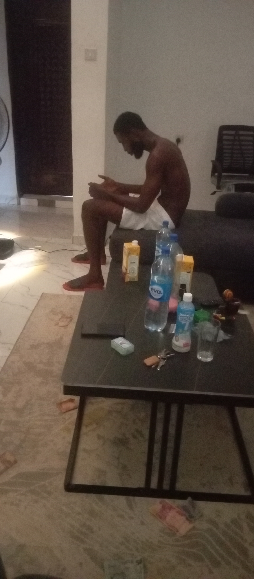 The image captures an indoor scene featuring a man sitting on a dark gray sofa, viewed in profile from the right. He is shirtless, wearing white shorts and red slide sandals, and has a beard. His head is slightly bowed, and he appears to be looking down at his hands, possibly engrossed in a mobile device or in thought.
In front of the sofa is a black coffee table cluttered with various items. These include several plastic water bottles, with at least two clearly labeled "Eva." Another water bottle has "Aqua Mist" partially visible. Two carton juice boxes are also present, one distinctly displaying "Hollandia Choco." A black smartphone, a set of keys, and a clear empty glass are arranged on the table. A small white rectangular box, possibly containing mints or gum, and a dark, decorative mortar and pestle are also visible. A remote control is partially obscured.
The room features light-colored walls and a light-toned tiled floor, partially covered by a beige patterned rug. Scattered across the rug and floor are several banknotes, one faintly showing the denomination "50." In the background, a dark, patterned door is visible to the left, and a dark office chair can be seen further back to the right. A portion of a floor fan is also visible on the far left. The lighting is soft and seems to be artificial, suggesting the scene takes place in the evening or night. The beverage brands and currency provide cues hinting at a West African, likely Nigerian, setting. The overall mood appears casual and relaxed.