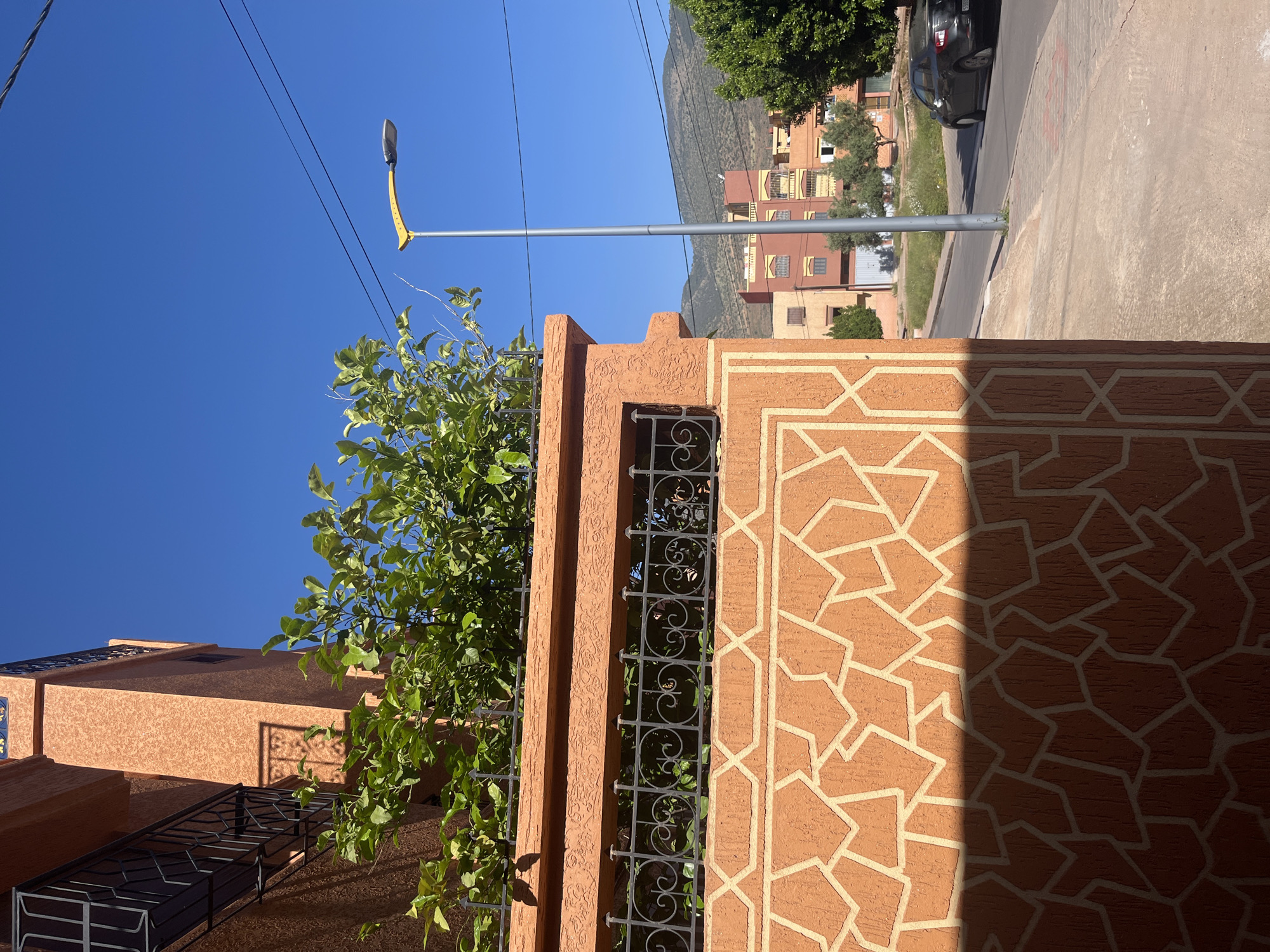The image shows a street scene in Ouaouizert, Morocco. A tall, terracotta-colored wall with geometric patterns and decorative ironwork runs diagonally across the frame. A tree with green leaves obscures part of the wall. A modern street light stands behind the wall, with power lines visible against a clear blue sky. In the background, multi-story buildings and a car parked along the side of the road can be seen. A mountain range is faintly visible in the distance. The ground is concrete. The scene appears to be brightly lit, with some areas of the wall in shadow.