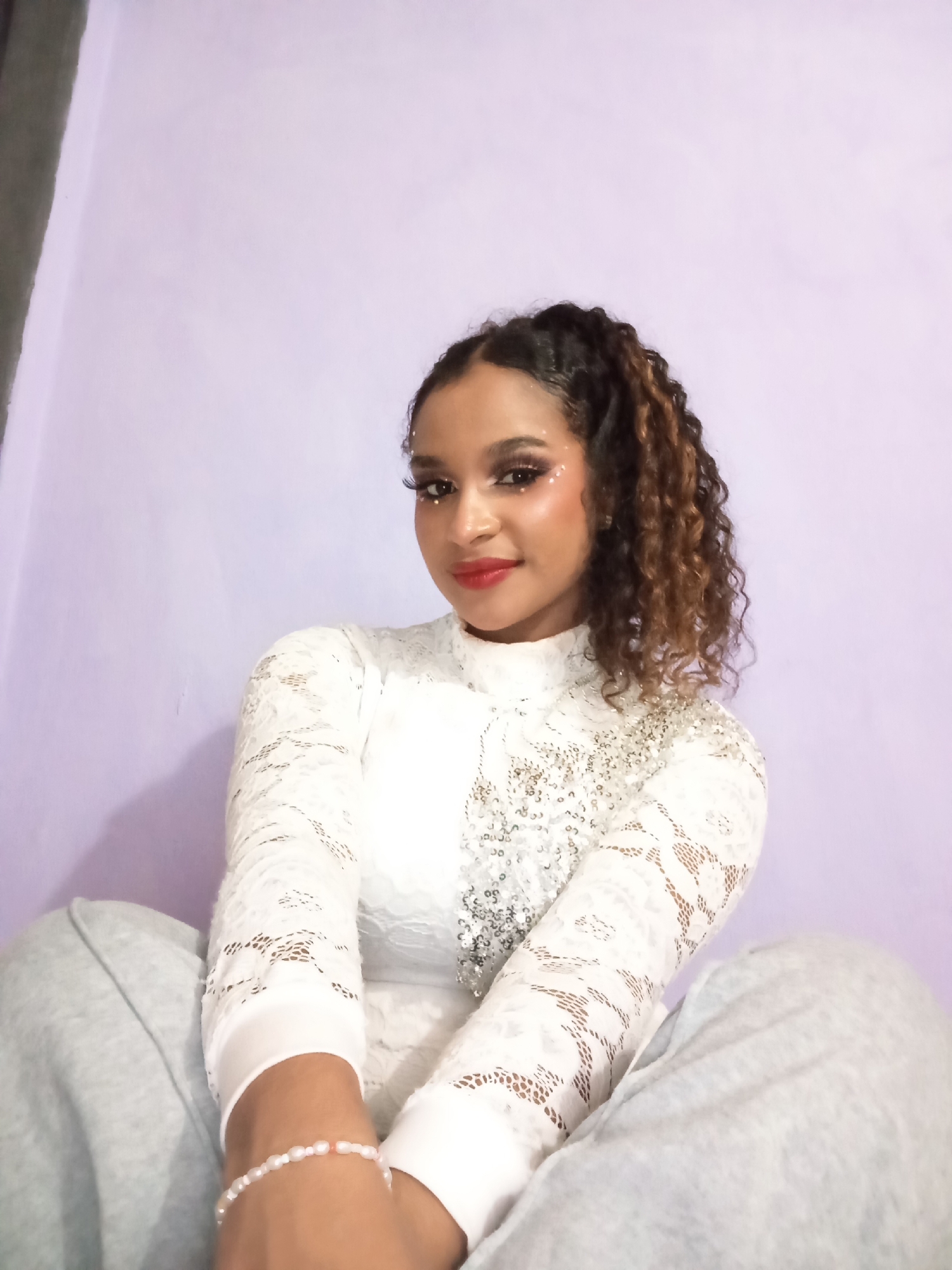 A young woman is sitting in front of a light purple wall. She is wearing a white lace top with long sleeves and silver sequins on the right side. She has her curly brown hair pulled back into a high ponytail. The woman is wearing makeup, including red lipstick and glitter around her eyes. She is wearing a pearl bracelet on her left wrist. The woman is sitting with her legs crossed, and she is wearing gray sweatpants. The photograph could have been taken in Juan Griego, Venezuela, or elsewhere.