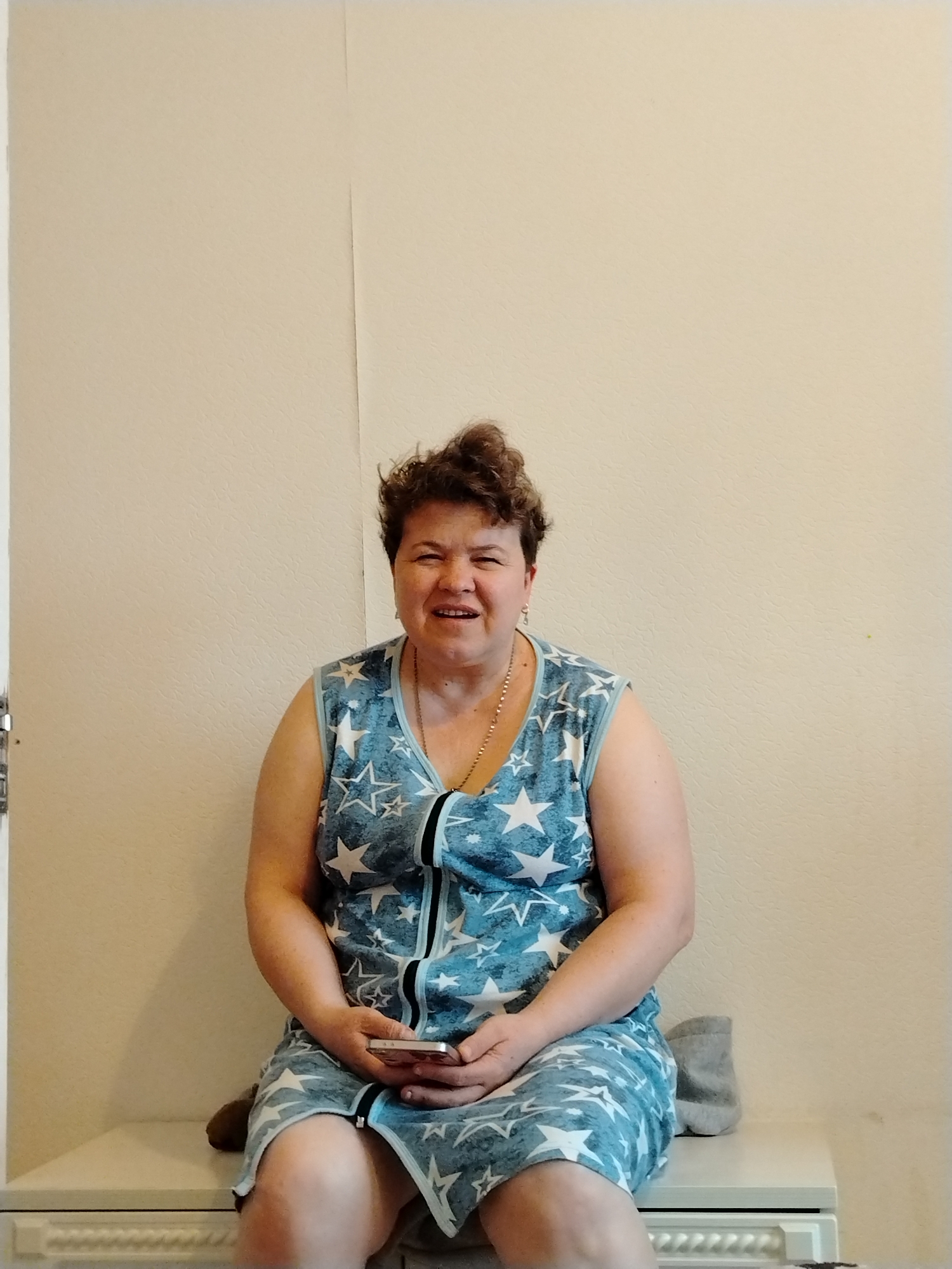 The image shows a middle-aged woman with short, curly brown hair sitting indoors. She is wearing a sleeveless, blue dress with white star patterns and a black zipper down the front. She is holding a smartphone in her hands. A gold necklace is visible around her neck. She is seated on a white or light-colored cabinet or bench. Behind her is a beige or off-white wall with a vertical seam or division. To the left of the woman, a white door frame is visible. To the right of the woman, is a sock. The image does not include enough details to confirm the location is in Noviy, Russia.
