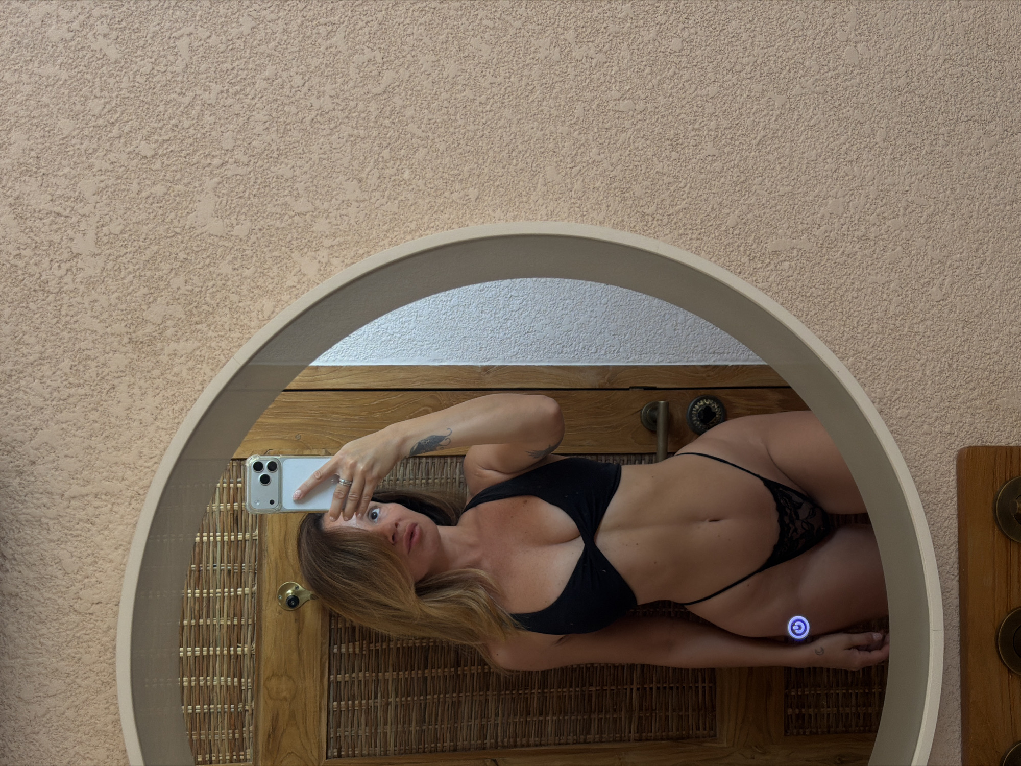 A woman in a black bikini is taking a mirror selfie in a bathroom. She has blonde hair and is wearing a ring on her left hand. The bathroom has a textured, beige wall and a wooden door. There is a woven basket pattern on the door. The mirror is oval-shaped and has a light-colored frame. A power button icon with a moon is visible on the woman's left wrist. The location is stated to be in the city of Mengwi, Indonesia.
