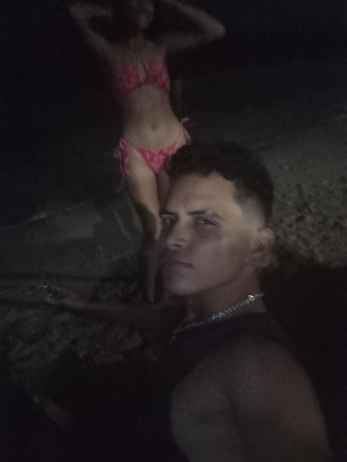 The image depicts two individuals at night in a coastal area of Juan Griego, Venezuela. In the immediate foreground, positioned on the right, is a male subject visible from the upper torso, wearing a dark garment and a silver-colored chain necklace. He has short, dark, curly hair with a faded cut on the sides, and his gaze is directed slightly left of the camera. In the mid-ground, standing further back and slightly to the left, is a female subject wearing a pink two-piece bikini. Her arms are raised above her head in a pose. She appears to be standing in shallow water or at the water's edge. The environment is dark, with indistinct ground texture in the foreground transitioning to a dark, reflective water surface. The overall lighting is low, creating a dimly lit scene.