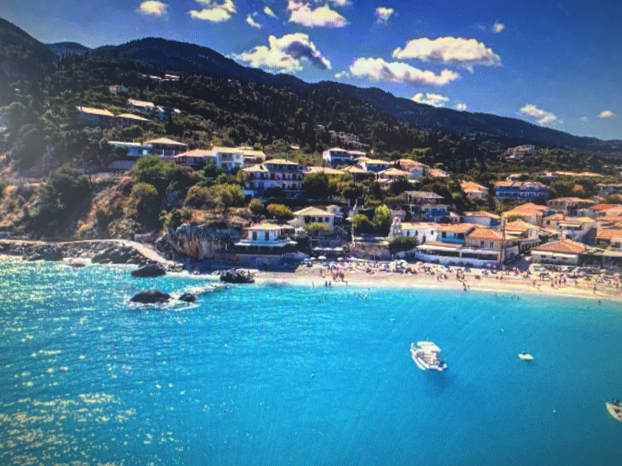 This media file captures a picturesque aerial view of a coastal village nestled at the foot of a lush, forested mountain. The scene is bathed in bright sunlight, suggesting a clear, warm day, likely midday or early afternoon, given the strong illumination and scattered white clouds against a vibrant blue sky.
The village itself is characterized by numerous buildings with terracotta-tiled roofs, clustered along the hillside and extending towards a sandy beach. These structures appear to be a mix of residential homes and possibly hotels or guesthouses, painted in light colors. The hillside is densely covered with green trees and shrubs, with some of the buildings seemingly built directly into the slope.
A busy beach is visible, dotted with numerous people enjoying the sunny weather. Some are sunbathing on the sand, while others are in the shallow water, engaged in various beach activities. The water is a striking, clear turquoise, with gentle waves lapping against the shore. A few small boats, including a white motorboat, are anchored or cruising in the bay, adding to the sense of a popular vacation destination.
The presence of rocky outcrops along the coastline and the overall Mediterranean-like appearance of the architecture and vegetation hint at a location in a region with a warm climate, possibly a European coastal area. The image evokes a feeling of leisure and relaxation associated with a summer holiday. No visible text is present in the media file.