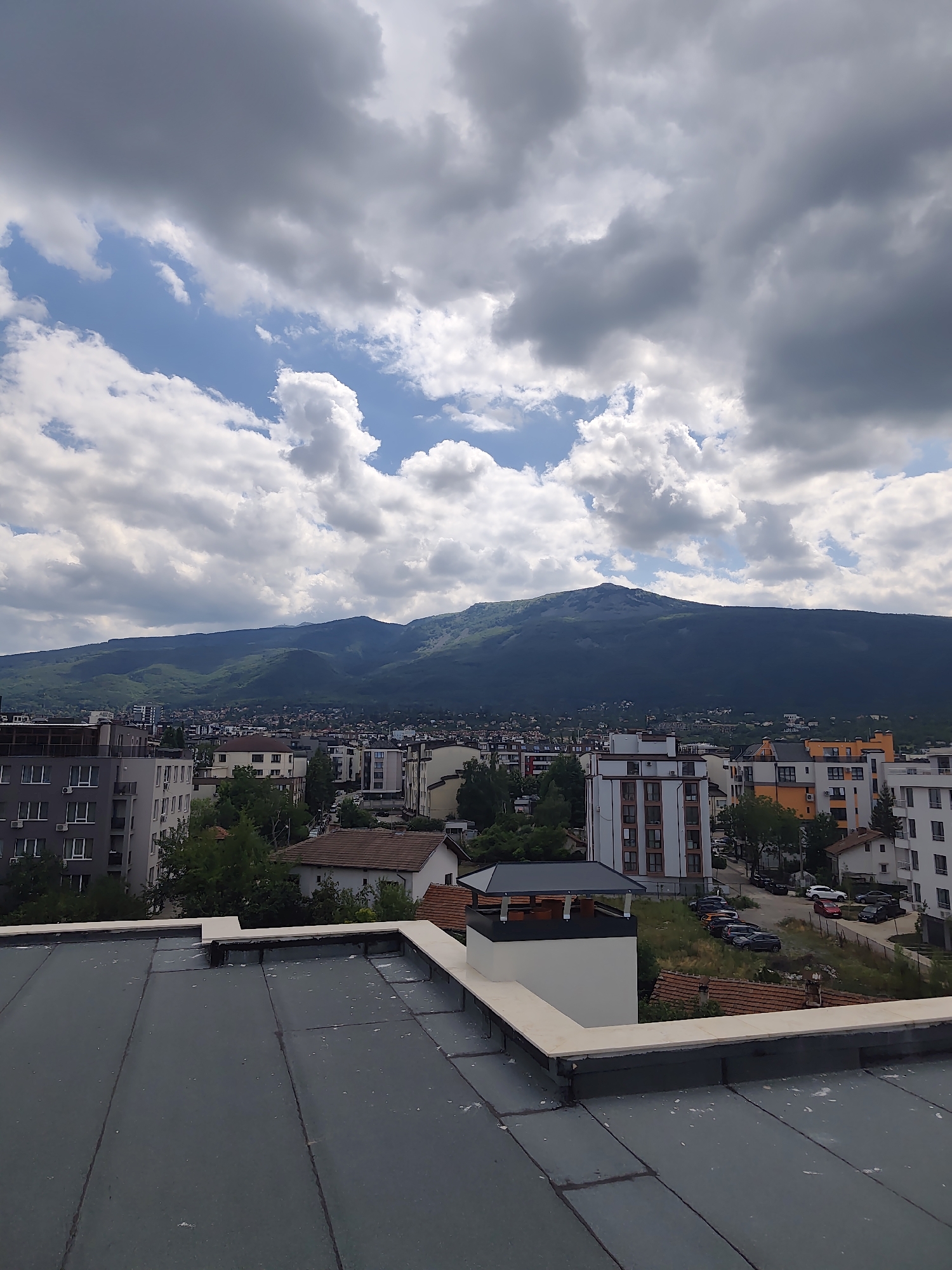 The image is an exterior, eye-level shot of a city skyline with a large mountain range in the background, taken from a rooftop. The foreground is dominated by a flat, dark grey rooftop with visible seams and some minor debris. Beyond the rooftop, a dense urban area with a mix of modern apartment buildings and older houses is visible. Trees and greenery are interspersed throughout the cityscape.
In the mid-ground, several multi-story buildings stand out, including a white and orange building with multiple balconies and a modern grey building with many windows. A parking lot with several cars is also visible between the buildings.
The background is dominated by a vast, green mountain range, shrouded in part by clouds. The sky above is a mix of blue patches and heavy, grey clouds, suggesting a dynamic weather system. The lighting is diffuse, indicating an overcast or partly cloudy day.
The scene appears to be in Sofia, Bulgaria, likely overlooking the Vitosha mountain range, which is a prominent natural landmark situated on the outskirts of the city. There are no people or animals visible in the frame.