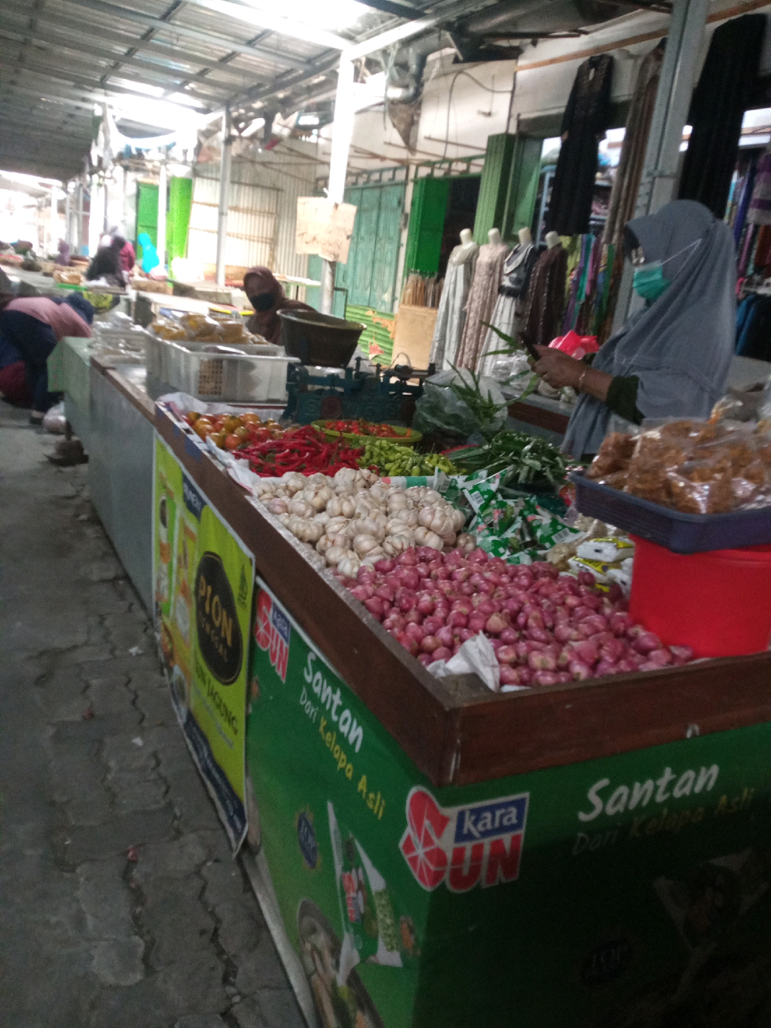 In Balun, Indonesia, a bustling market scene unfolds, captured from a low angle. The foreground is dominated by a display of fresh produce, with piles of shallots and garlic neatly arranged. Behind them, a vibrant assortment of red and green chilies, along with tomatoes, adds a splash of color. A woman, partially obscured by the produce, wears a grey hijab and a light green face mask, holding a smartphone in her hands. She stands behind a stall displaying various packaged goods, with a red plastic container and a dark woven basket filled with fried snacks on the counter. The market stall features green banners with Indonesian text, including "Santan Dari Kelapa Asli" (Coconut Milk from Original Coconut) and "PION JAGUNG" (Corn PION). In the background, mannequins are dressed in traditional Indonesian attire, and other vendors and shoppers are visible, suggesting a lively commercial atmosphere. The market is covered by a metal roof, typical of many Indonesian marketplaces. The ground is paved with cobblestones, showing signs of wear and tear.