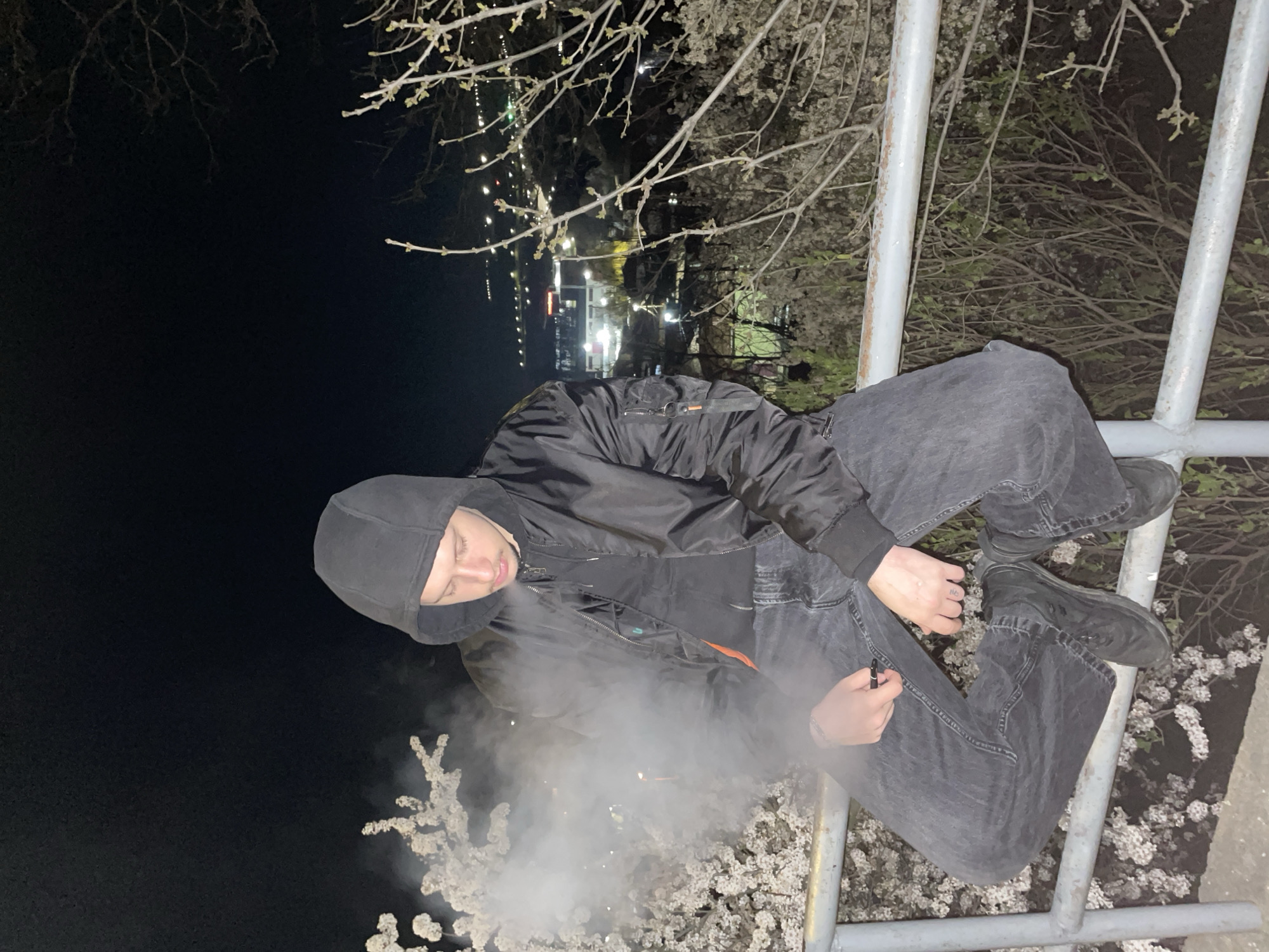 A young individual wearing a dark hooded jacket and dark jeans is perched on a gray metal structure, possibly a fence or railing, at night. The individual is exhaling a cloud of white vapor, possibly smoke from a vaping device or cigarette. The background consists of dark foliage, including blooming white flowering branches near the bottom of the frame. In the distance, there are out-of-focus lights suggesting a city or town at night. The location is Razgrad, Bulgaria.