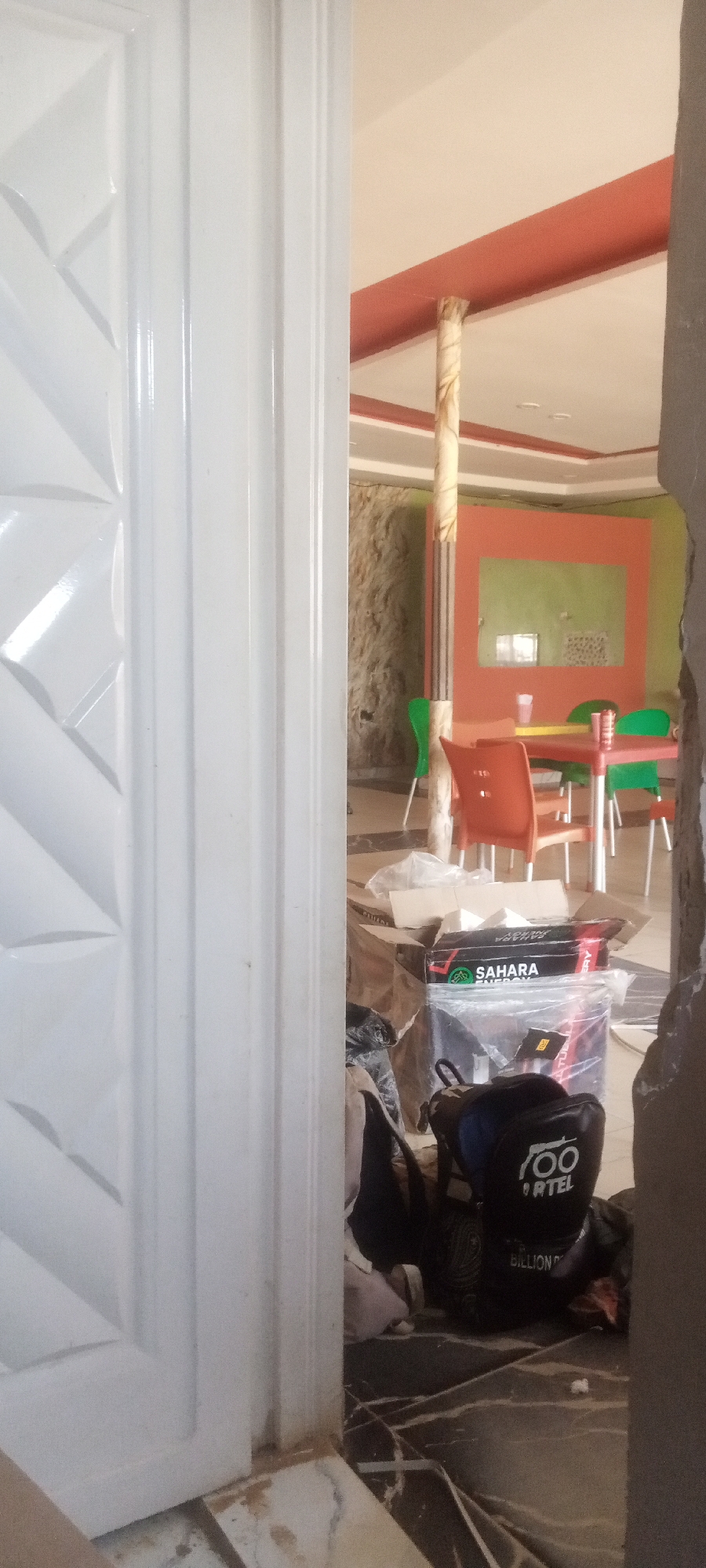 The image, taken in Sabon Gari Nassarawa, Nigeria, shows a partial view into what appears to be a dining area through a partially open white door. In the dining area, there are chairs and tables with a decorative partition in the background. Foreground details include a box labeled "SAHARA ENERGY", a bag with the word "ORTEL" printed on it, and general clutter on the floor. The floor is dark marble. The setting suggests ongoing renovation or setup of a commercial space, potentially a restaurant or cafe. There are no people visible in the frame.