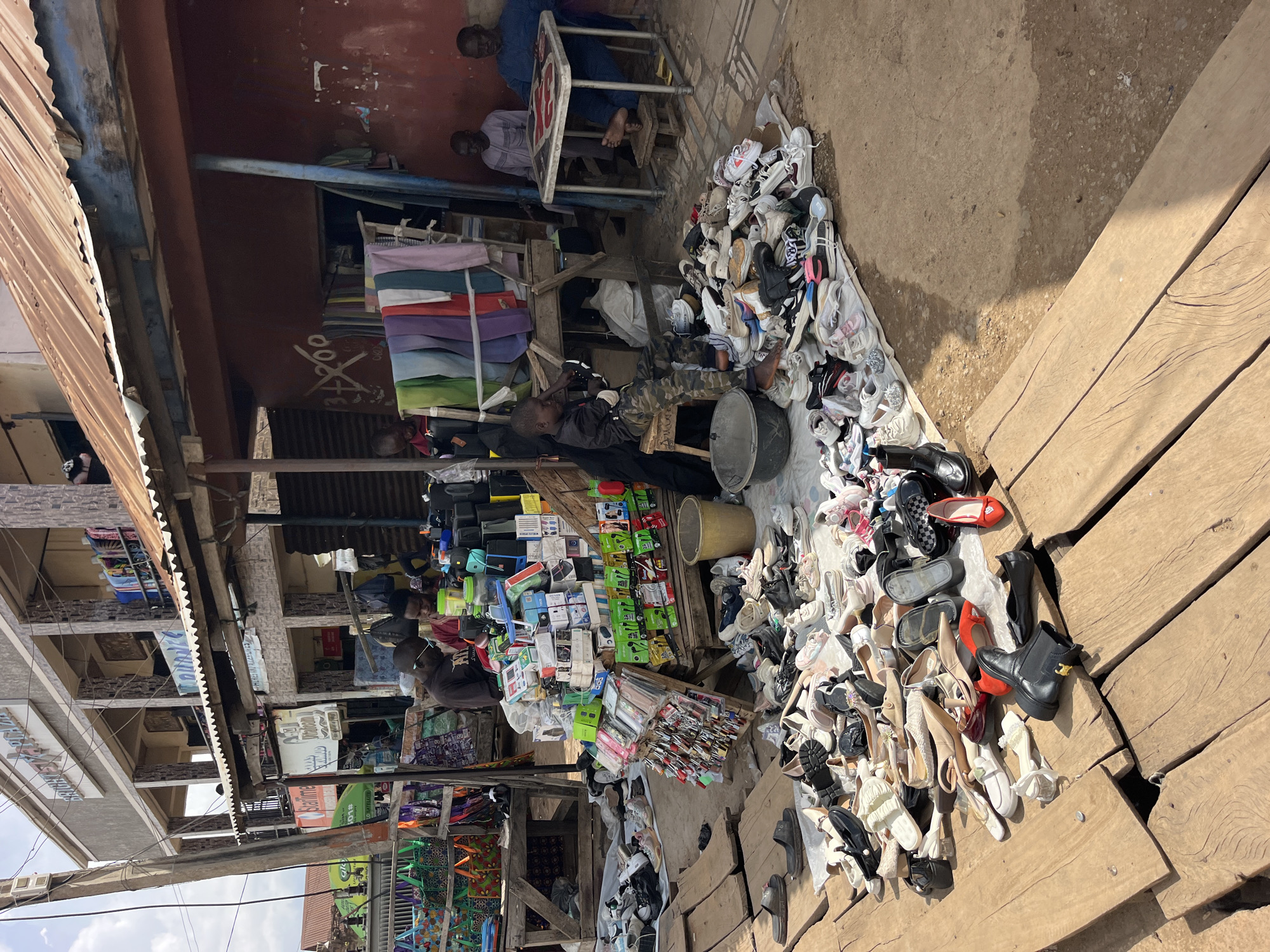 An outdoor market scene unfolds in Unknown, Unknown. A stall displaying an assortment of shoes is in the foreground, with various pairs of sneakers, sandals, and heels scattered across wooden planks and a light-colored tarp. To the right, a pile of shoes sits on the dirt ground. Behind the shoe display, a stall is laden with boxes of electronics, including cell phones and accessories, and a selection of colorful clothes hangs on a rack. Two men are seated at a table to the right, engaged in conversation. A young boy in camouflage pants sits on a stool, examining an item. In the background, a multi-story building with balconies and shops is visible. The scene is illuminated by daylight, casting shadows across the ground.
