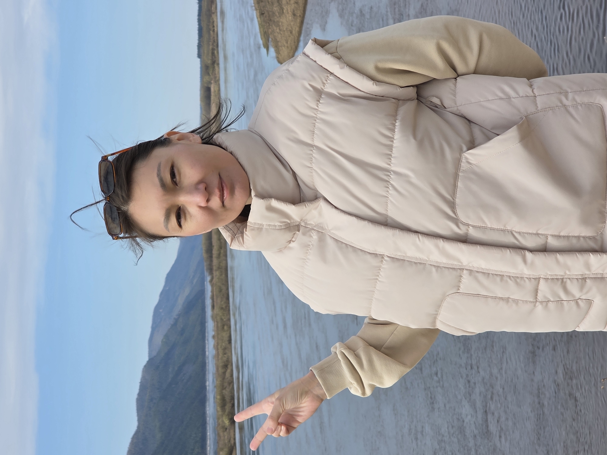 The image shows a woman standing outside, likely in Lensk, Russia, with a river and landscape in the background. The woman is wearing a light-colored quilted vest over a tan sweatshirt. Sunglasses are perched on her head. She is making a peace sign with her hand. The background consists of a wide river, a distant shoreline with vegetation, and hills under a blue sky.
