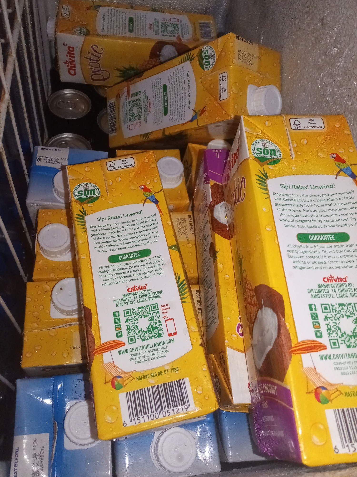 The image shows a close-up, interior view of a refrigerator filled with various cartons of Chivita Exotic fruit juice. The cartons are predominantly yellow and feature fruit imagery, including pineapples and coconuts, as well as parrots. Some cartons also display the "SON" logo and "FSC" certification. Several of the cartons have white plastic caps. The lighting is dim, suggesting the refrigerator is on. There are no people or animals visible in the image. The scene appears to be a commercial or domestic refrigerator unit.