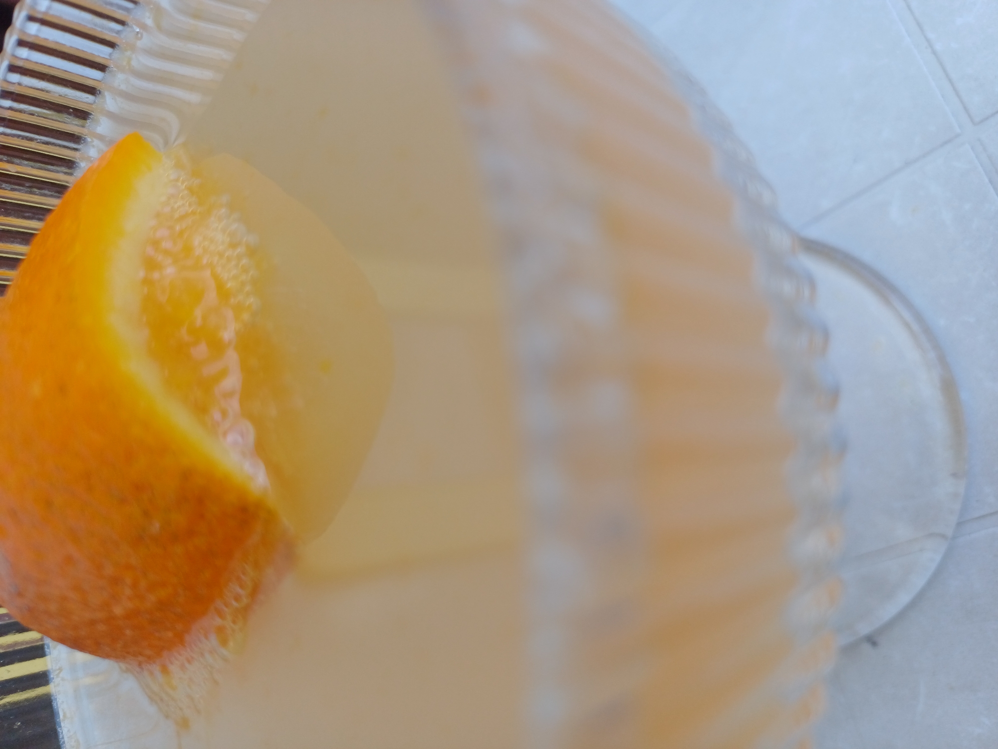 A close-up, slightly out-of-focus shot captures a segment of a clear, fluted glass bowl filled with a pale orange liquid. A wedge of orange peel, with its textured rind and pith, is submerged in the liquid, creating a small ring of bubbles around its edges. The bowl sits on a white surface, possibly a table, with faint lines suggesting tiles or a patterned cloth underneath. The background is blurred, with vertical stripes of black and yellow visible at the top. The scene is indoors, likely in a residential or hospitality setting. There are no individuals present in the frame. The setting is not identifiable as being in Varna, Bulgaria.